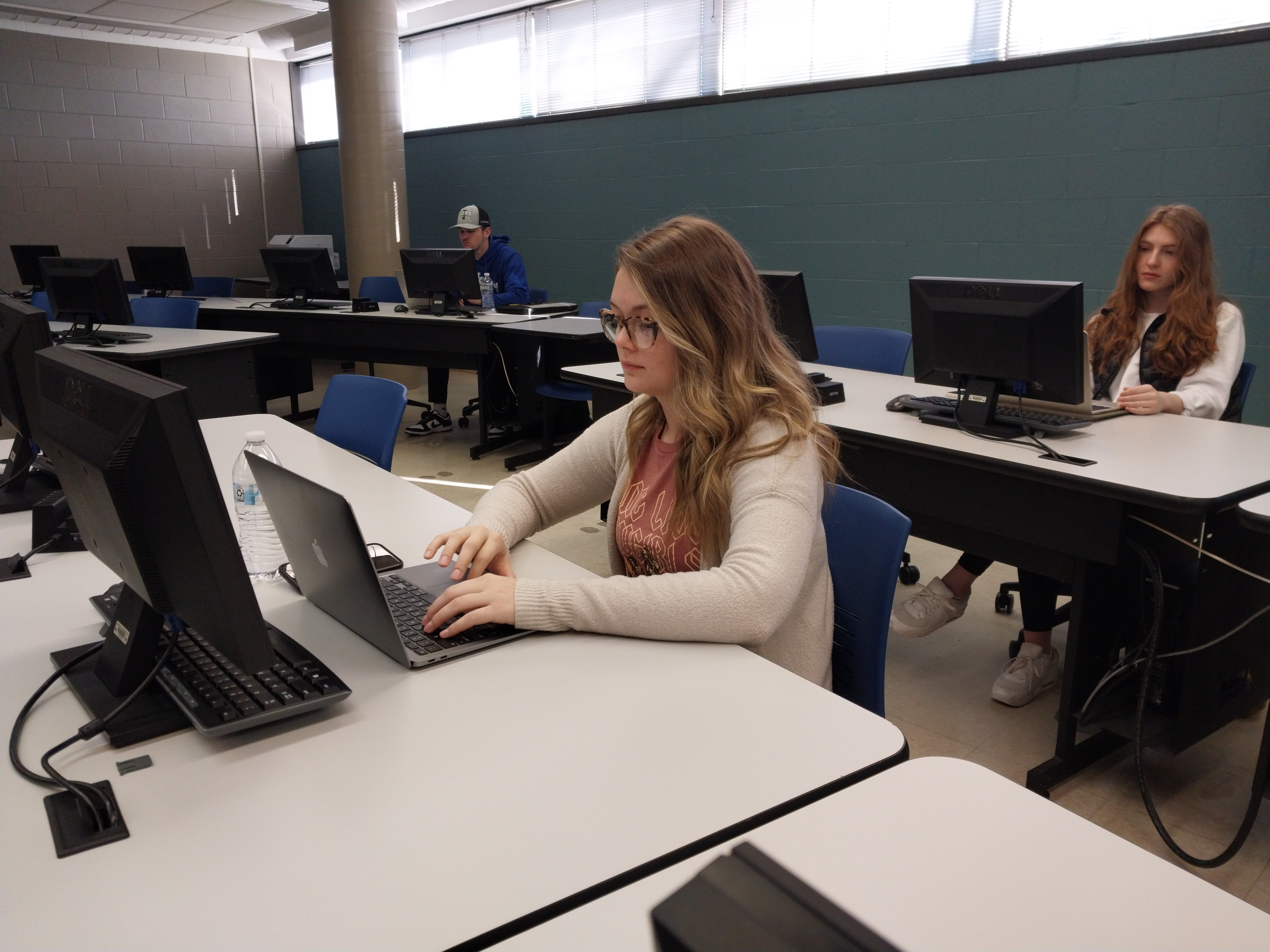 Student works on a laptop in a computer classroom while other students use desktop computers at individual workstations during an accounting or business course.