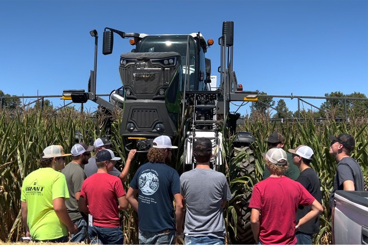 A group of students stands in a cornfield during an agriculture demonstration, facing a large self-propelled crop sprayer with its booms extended across the field. The students are gathered near the front tire and cab of the machine as they listen and observe on a clear, sunny day.