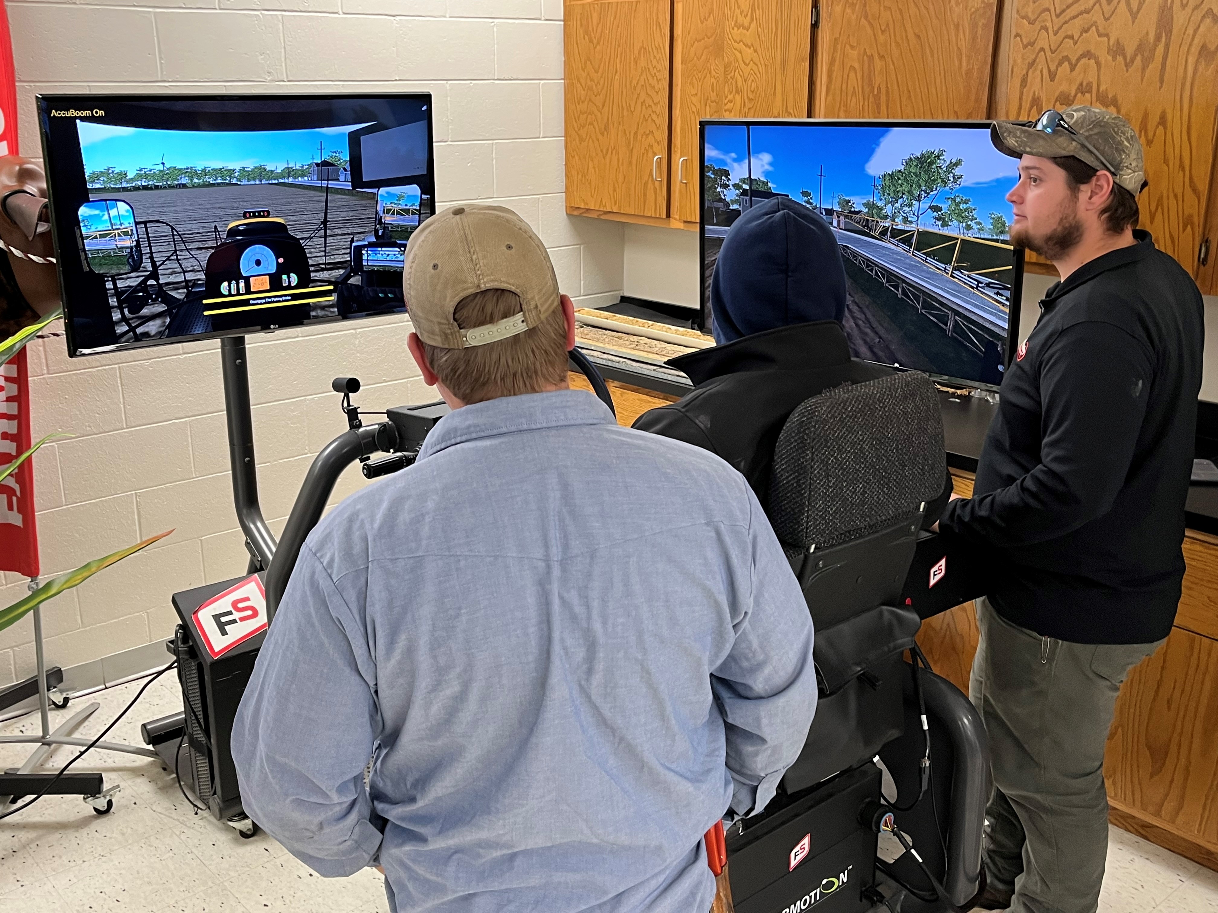 A student uses an agricultural equipment simulator while another student and an instructor observe nearby. Two monitors show a virtual view from inside a crop sprayer, including field rows, boom controls and guidance screens. The setup is in a classroom or lab space with cabinets and instructional equipment around the simulator.