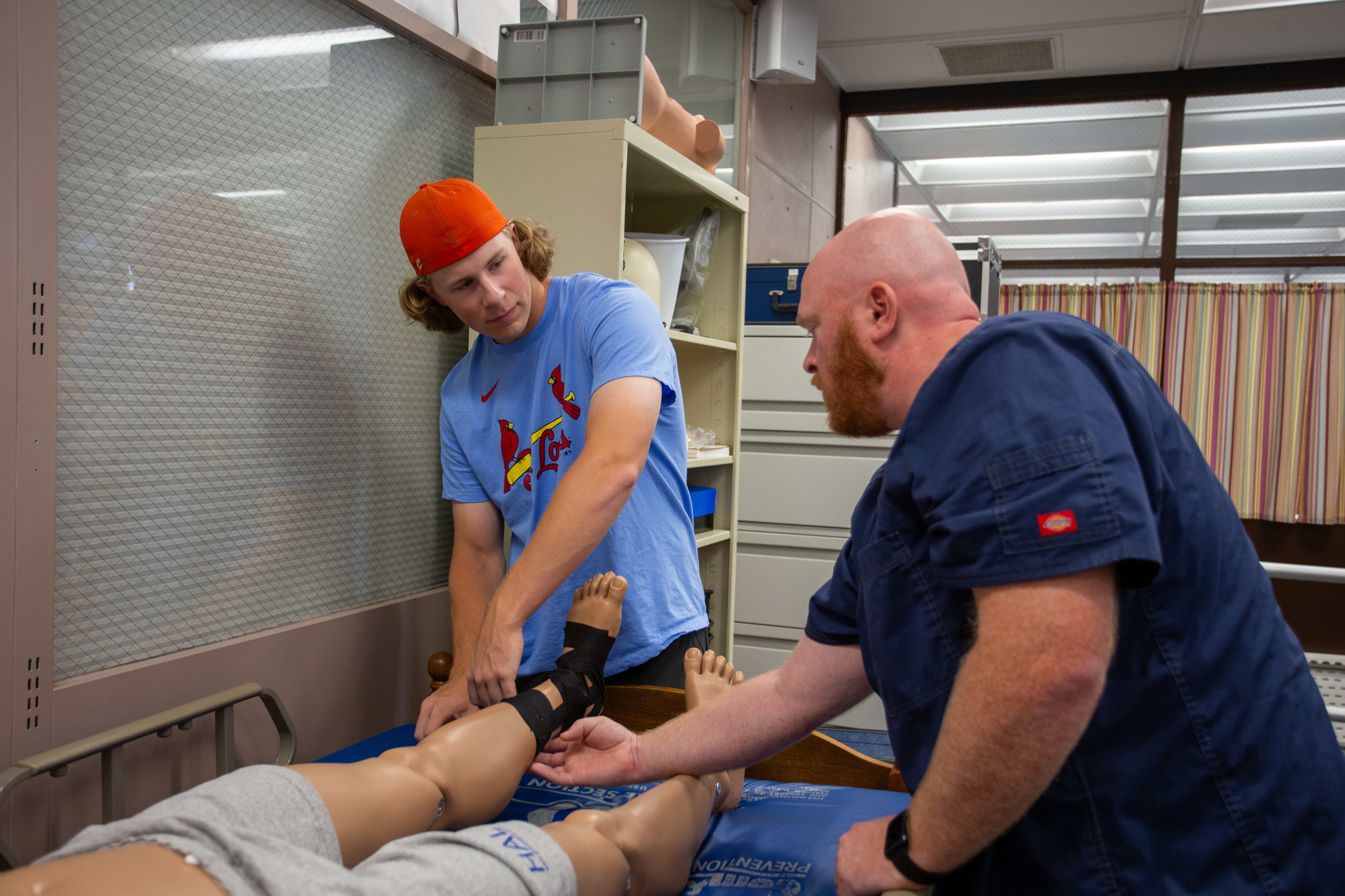 A student practices wrapping a mannequin’s ankle while an instructor watches closely in a clinical lab. The mannequin is lying on an exam table, and the student appears to be applying or adjusting athletic tape or a support wrap around the foot and ankle. Medical training equipment and storage cabinets are visible in the background.