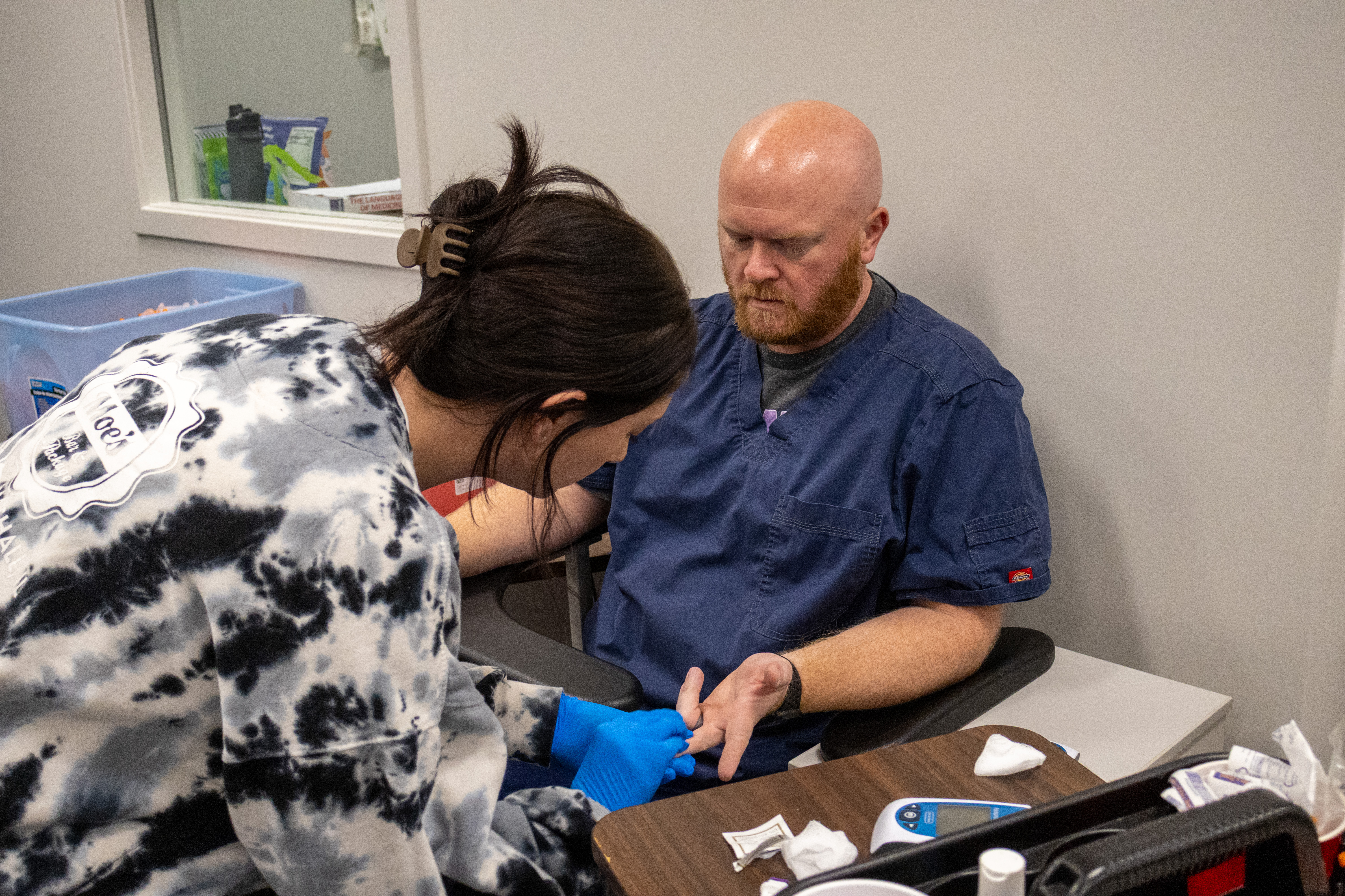 A student wearing gloves practices a finger-stick procedure on an instructor in a clinical lab. The instructor, wearing medical scrubs, sits beside a table with gauze, testing supplies and a small digital medical device while watching the student complete the hands-on training activity.