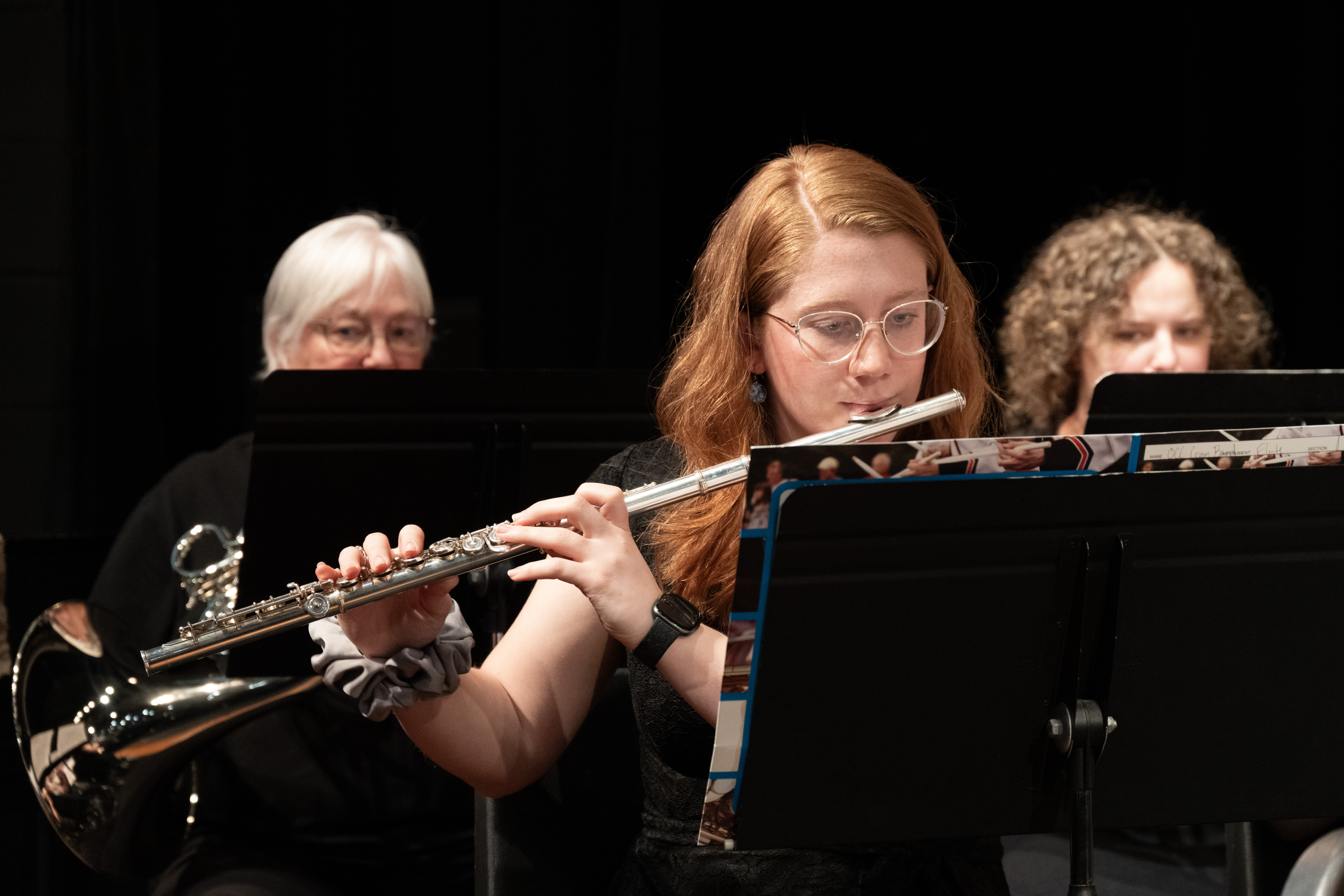 Student musician plays a flute during an ensemble performance on stage, reading sheet music from a stand. Other musicians and instruments are visible in the background.