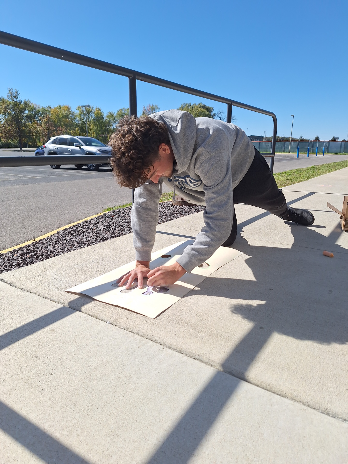 A student in a gray hoodie creates chalk art on a piece of cardboard laid on a sidewalk outside. He is leaning over the drawing and working with his hands on a sunny day, with a roadway, parked car, trees and blue sky in the background.