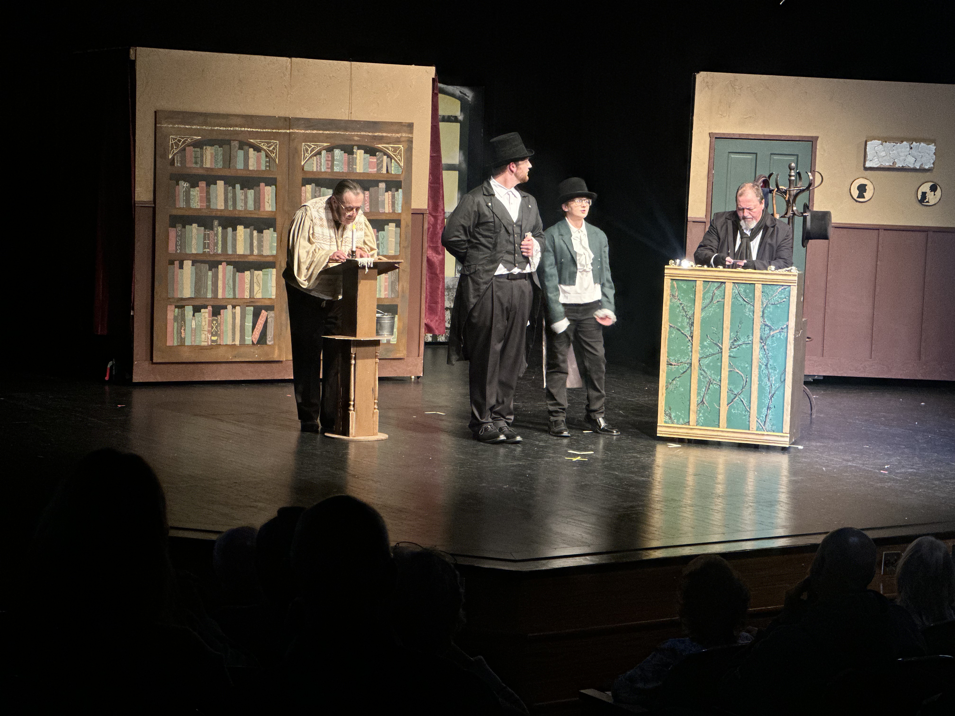 Four actors perform on stage in a theater production, wearing period-style costumes and hats. The set includes painted bookshelves, interior walls, a door and a small counter or desk. One actor writes at a small podium while three others stand nearby, including one actor positioned behind the counter. Audience members are visible in silhouette in the foreground.