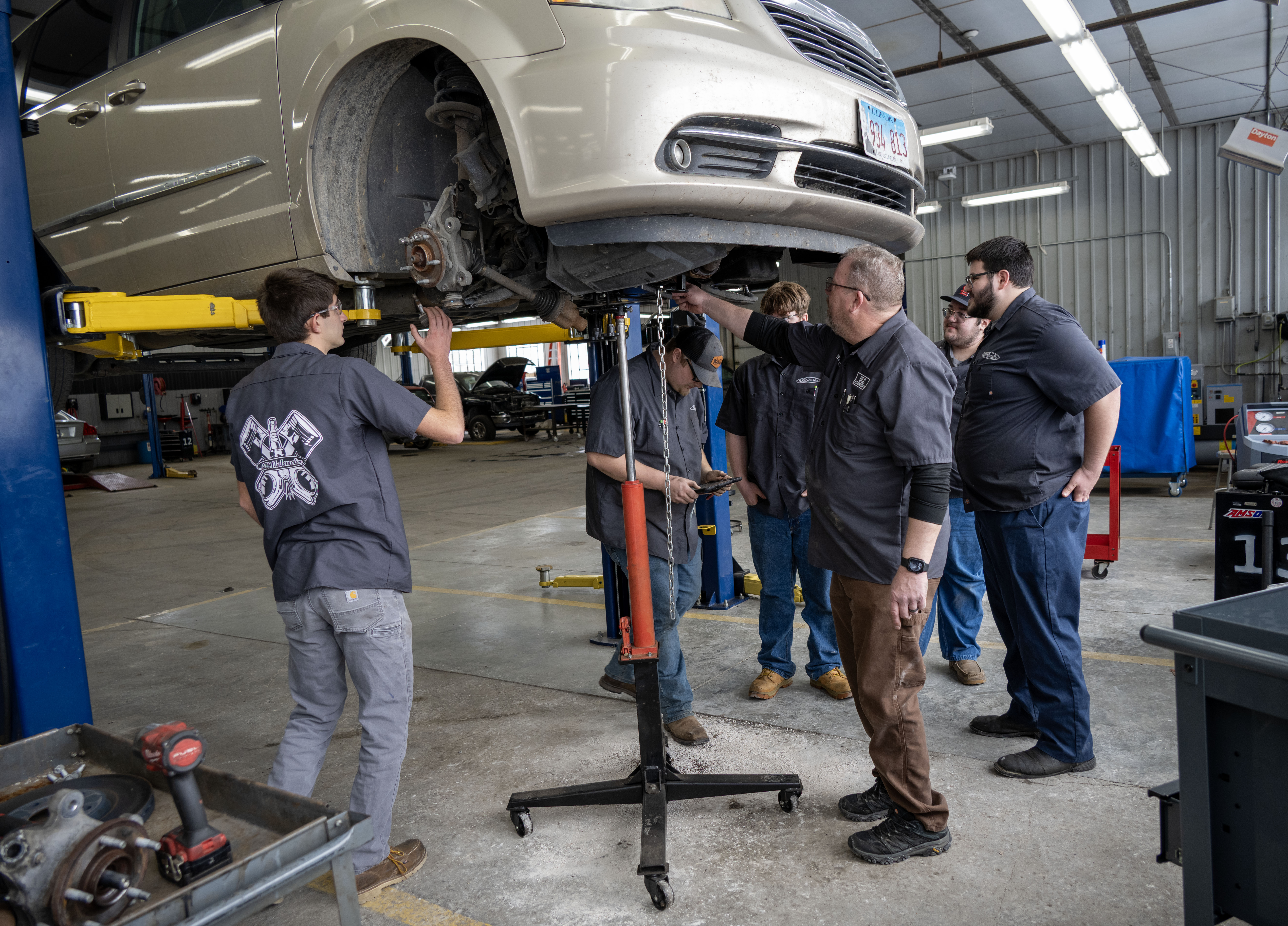 Automotive technology students and their instructor inspect the underside of a vehicle raised on a lift, performing diagnostic and maintenance procedures in a service lab.