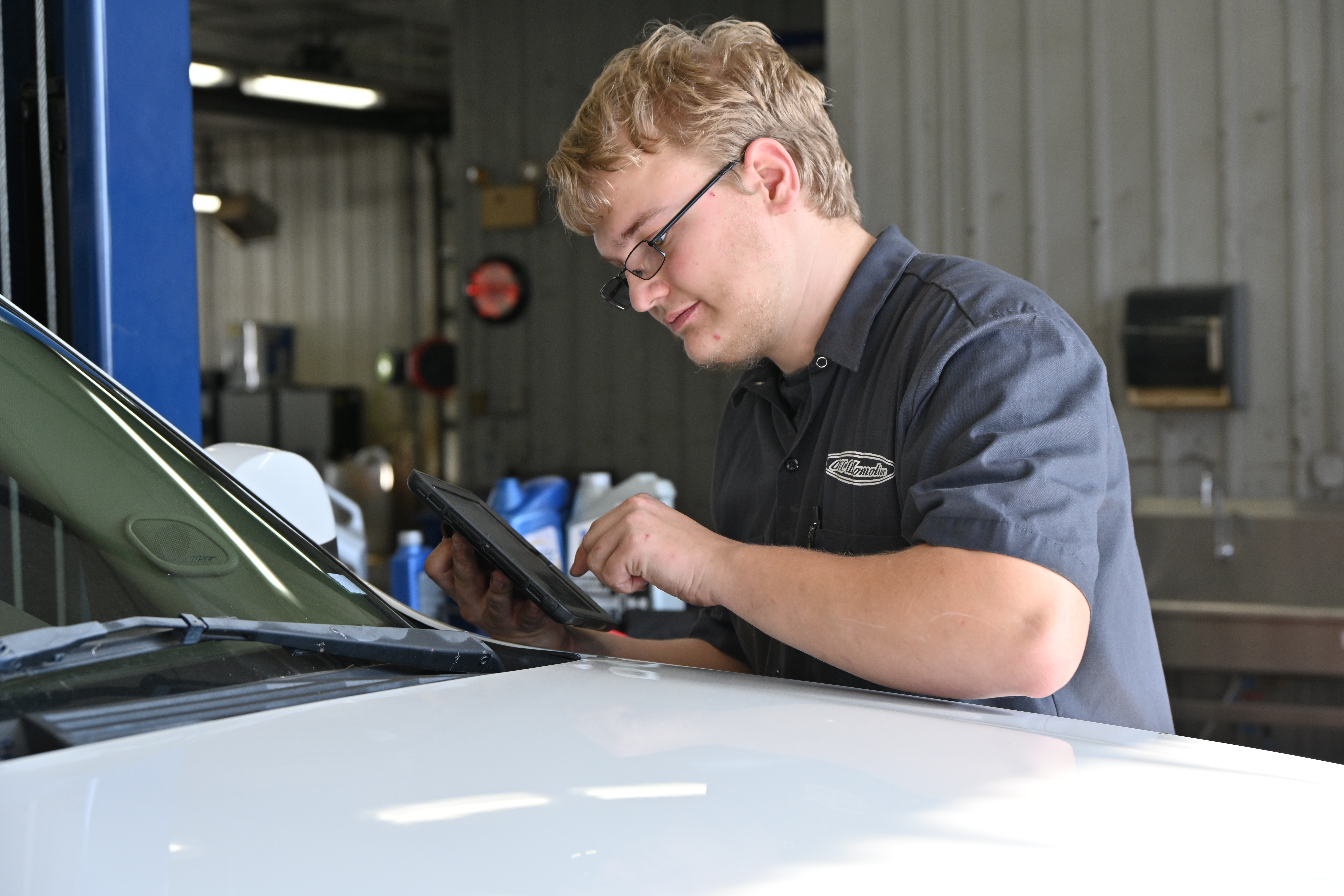 Automotive technology student uses a diagnostic tablet to inspect and troubleshoot a vehicle in a service lab, performing electronic diagnostics as part of hands-on automotive training.