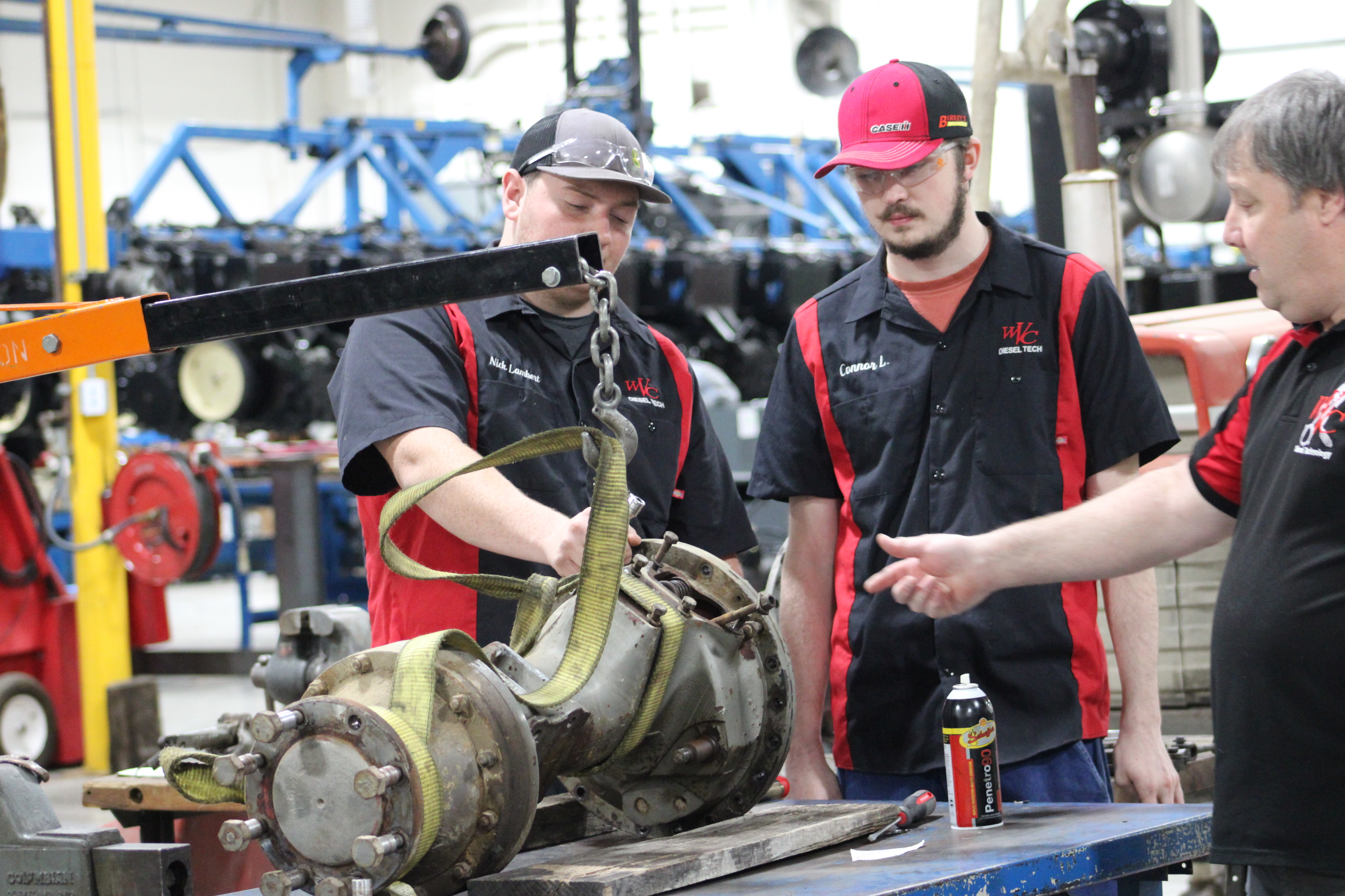 Two diesel technology students watch as an instructor points to a large mechanical component suspended from an engine hoist on a workbench. One student works near the component while the other observes. The scene takes place in a shop lab with tools, equipment and machinery visible in the background.