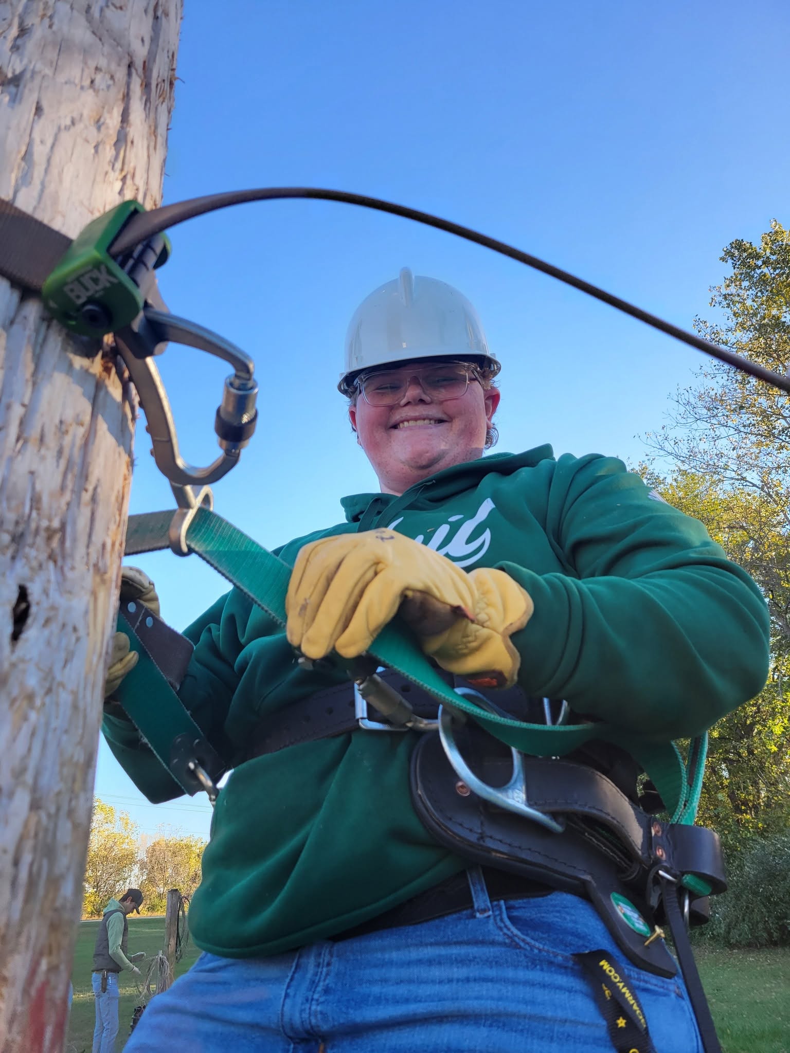 A broadband telecommunications student wearing a hard hat, safety glasses, gloves and a climbing harness smiles while secured to a utility pole. Safety straps, clips and cables are attached near the pole as part of a hands-on training exercise. Another person stands in the background near additional equipment on a clear day.