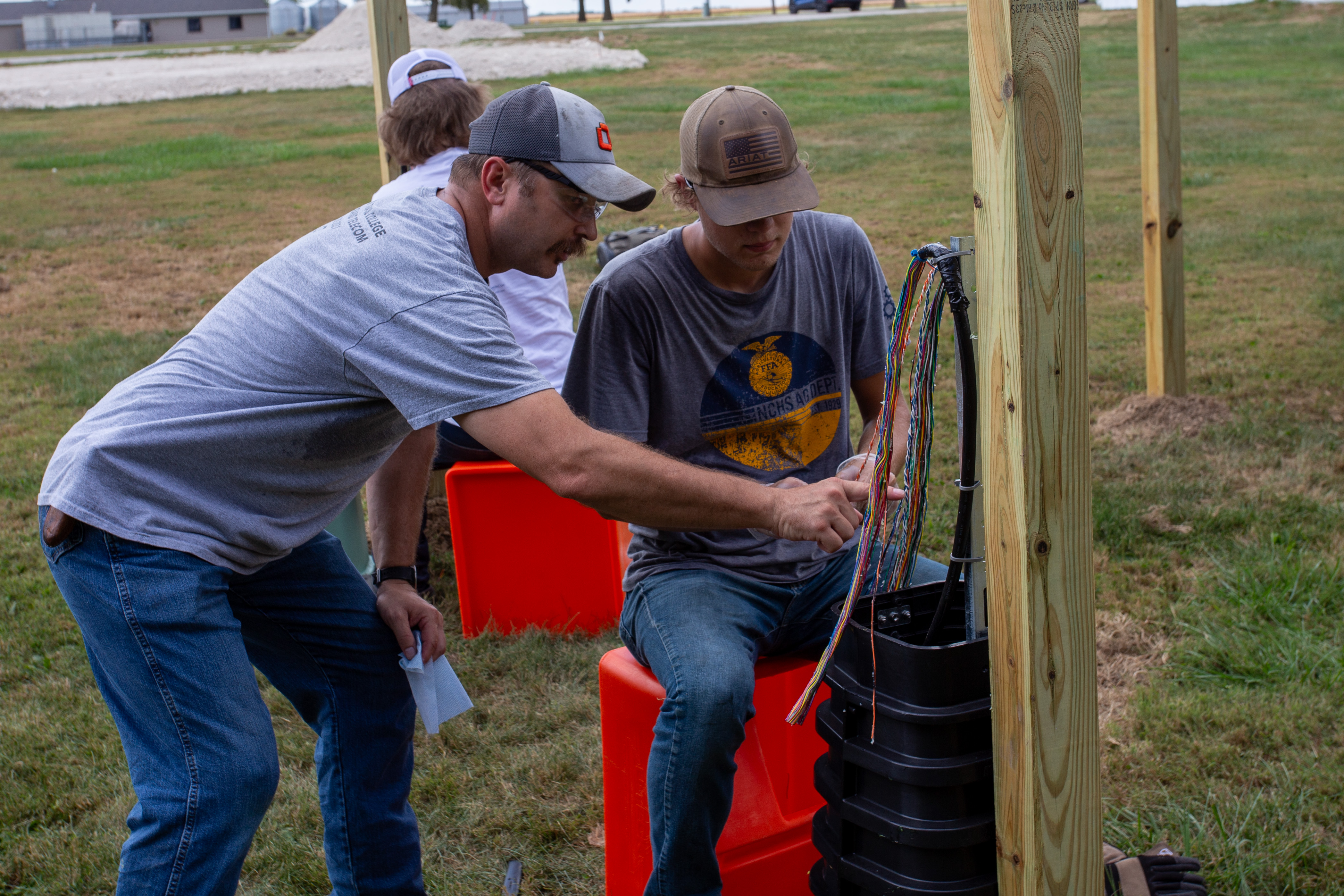 An instructor points to exposed colored wires inside a telecommunications service box mounted beside wooden training poles. A student sits nearby and studies the wiring during an outdoor broadband telecommunications lab, with other students and equipment visible in the background.