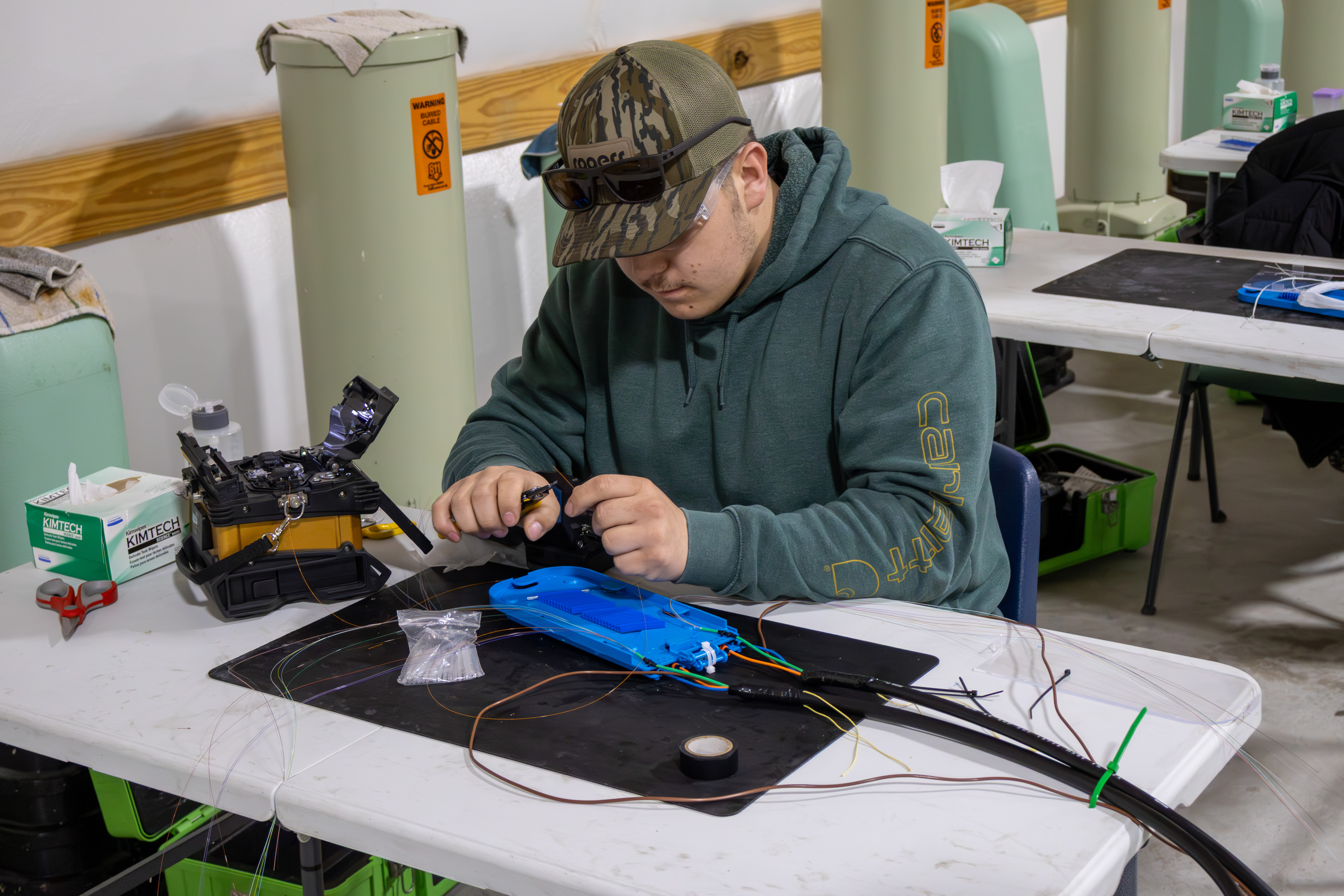 A broadband telecommunications student works at a table on fiber optic wiring and cable components during a hands-on lab. The student wears safety glasses and uses small tools while surrounded by colored wires, fiber splicing equipment, wipes and training materials. Utility pedestals and additional workstations are visible in the background.