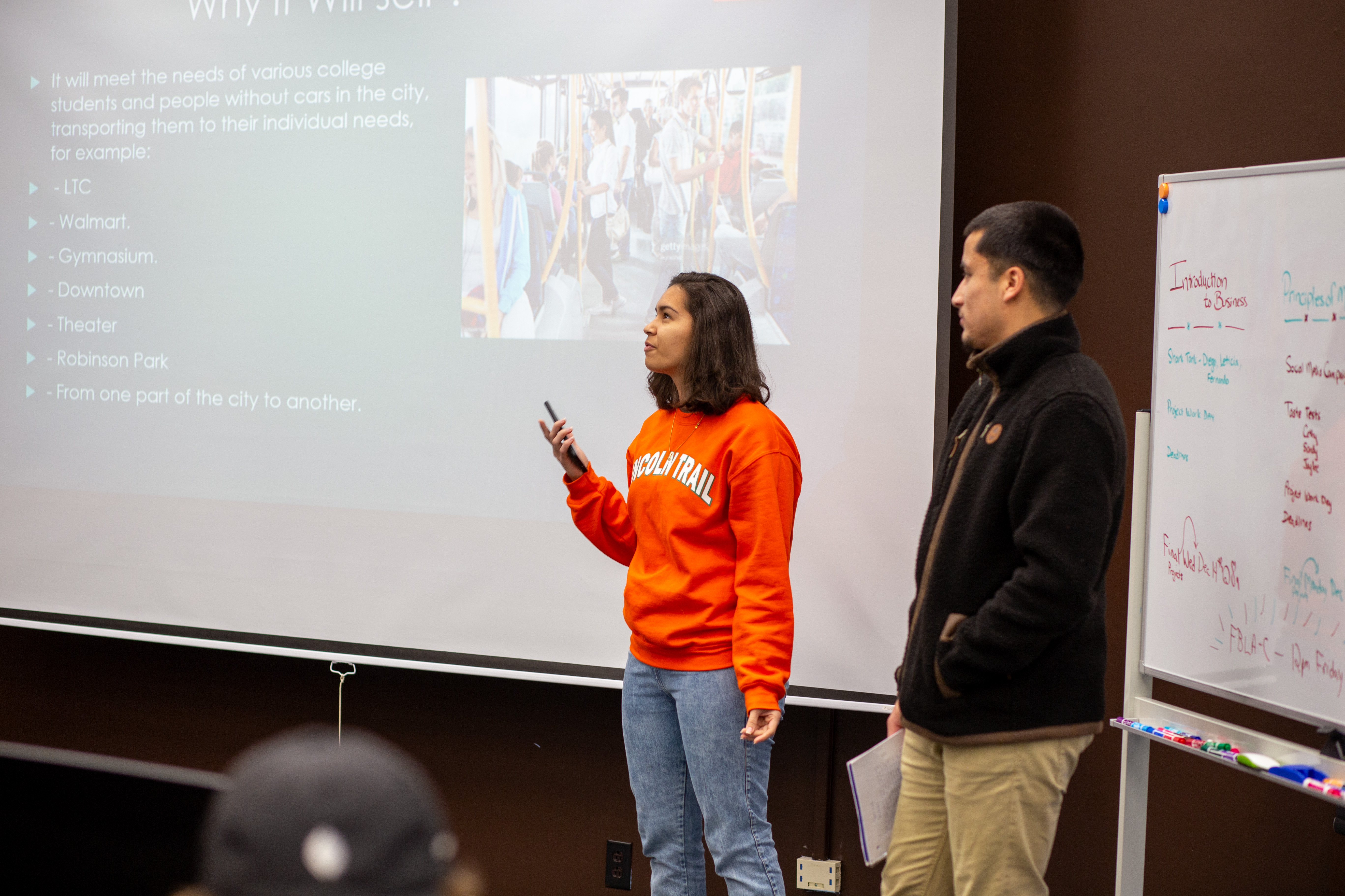 Two students give a business class presentation beside a projected slide about a proposed transportation service. One student speaks while holding a presentation remote, and another stands nearby with notes. The slide lists possible destinations, including LTC, Walmart, the gymnasium, downtown, the theater and Robinson Park. A whiteboard with class notes is visible beside them.