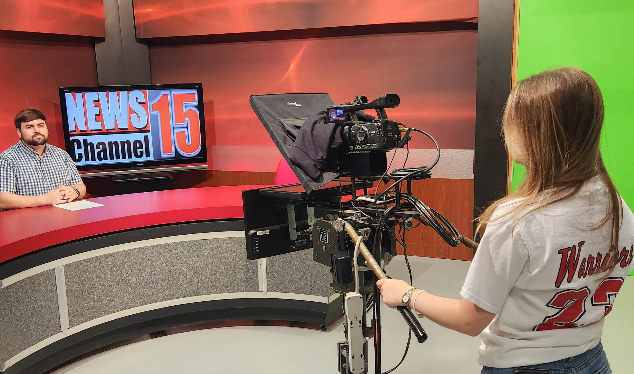 A student operates a professional television studio camera while another student sits at a news desk in front of a “News Channel 15” display. A green screen, studio lighting, teleprompter equipment and broadcast set pieces are visible, suggesting hands-on communications training in video production, news reporting and media broadcasting.