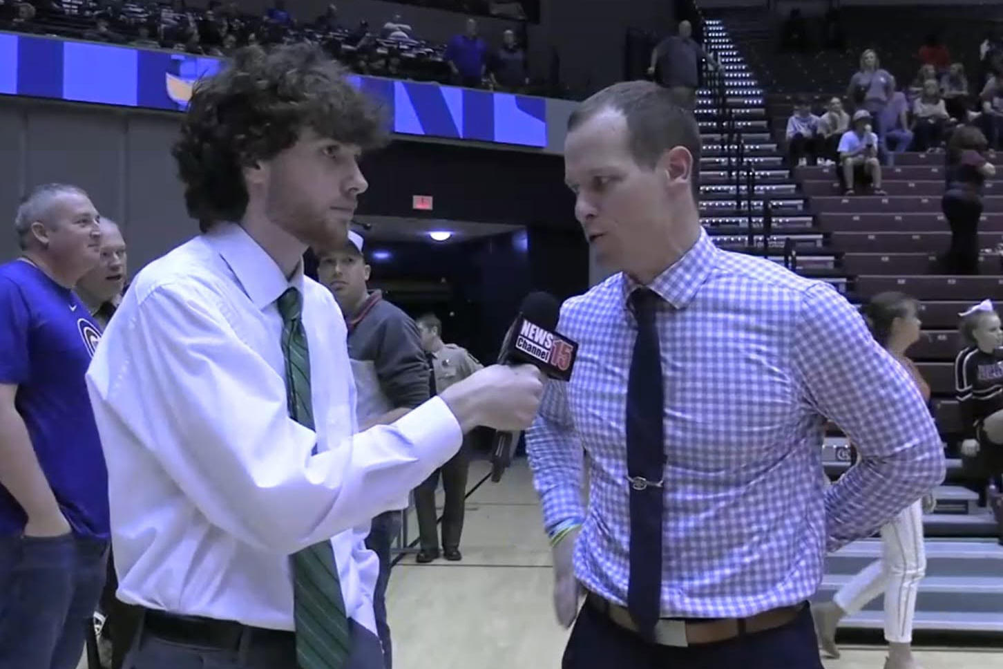 A student reporter holds a “News Channel 15” microphone while interviewing a coach or speaker courtside in a gymnasium after a game. Spectators and arena seating are visible in the background, emphasizing hands-on journalism experience in sports reporting, interviewing and live event coverage.