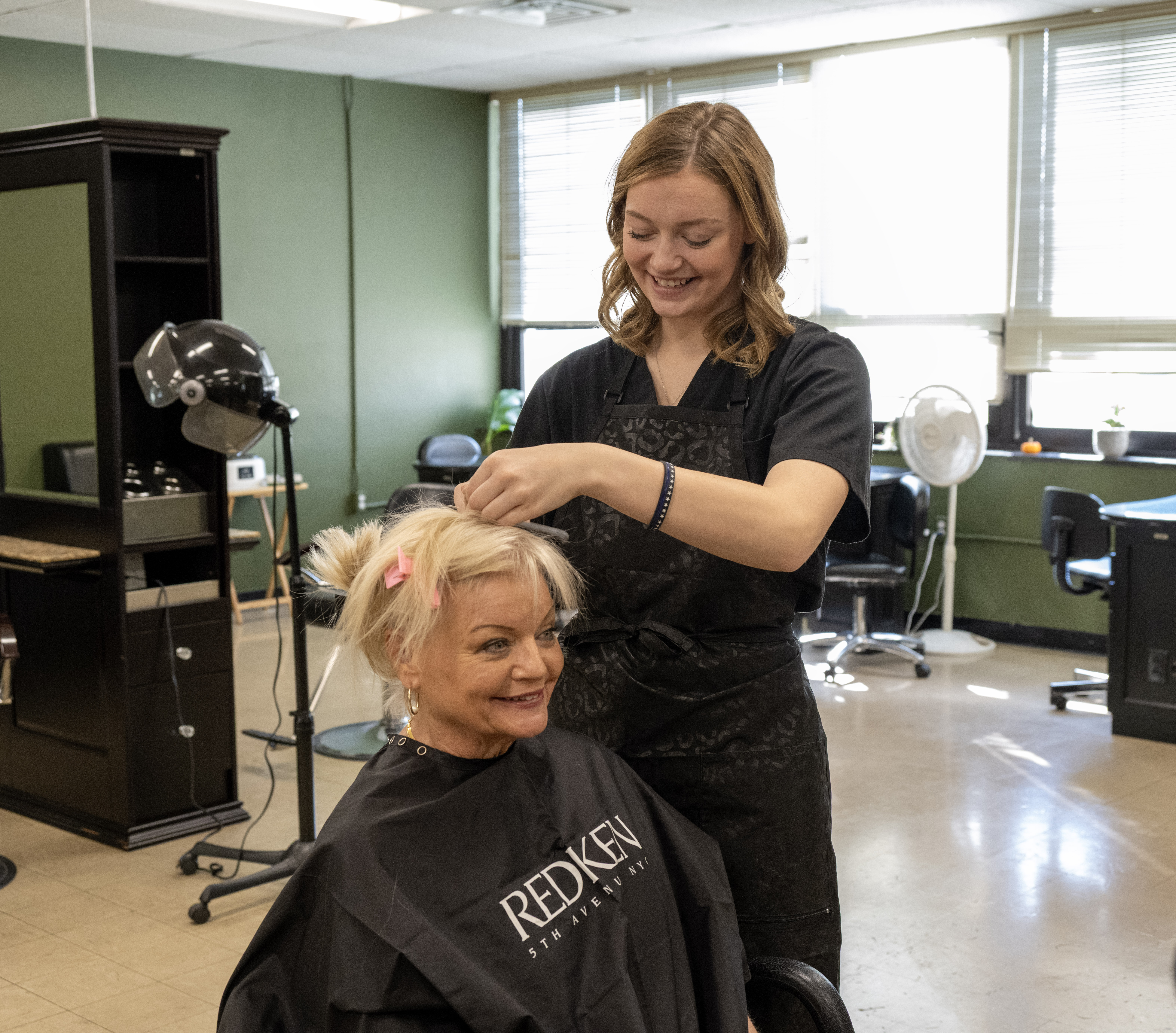 A cosmetology student practices hairstyling techniques on a seated client in a salon training classroom. Styling equipment and salon stations are visible in the background.