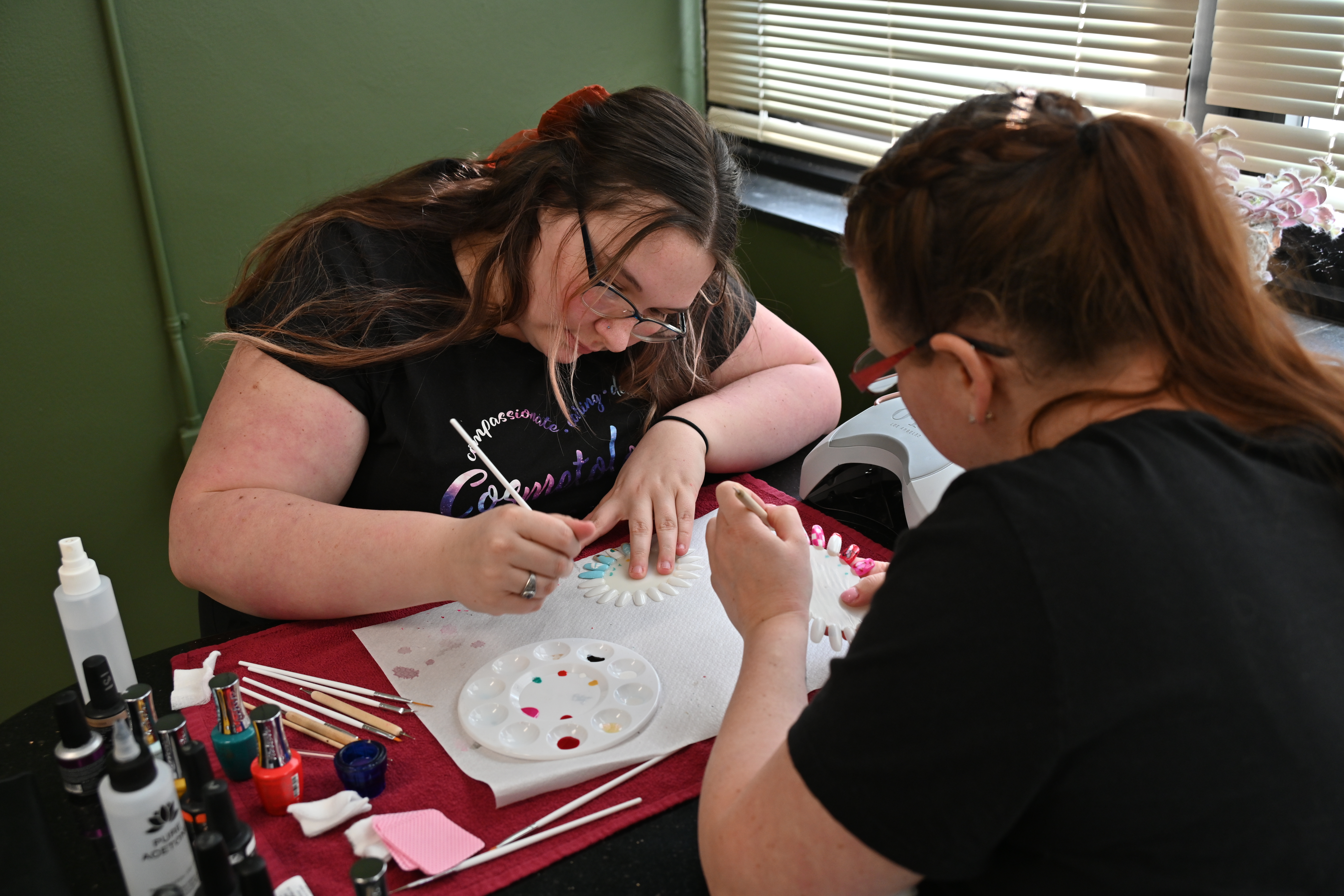 Two students work at a manicure station, painting and practicing nail designs on sample nail displays. Nail polish bottles, brushes, and training tools are arranged on the tabletop in the salon.