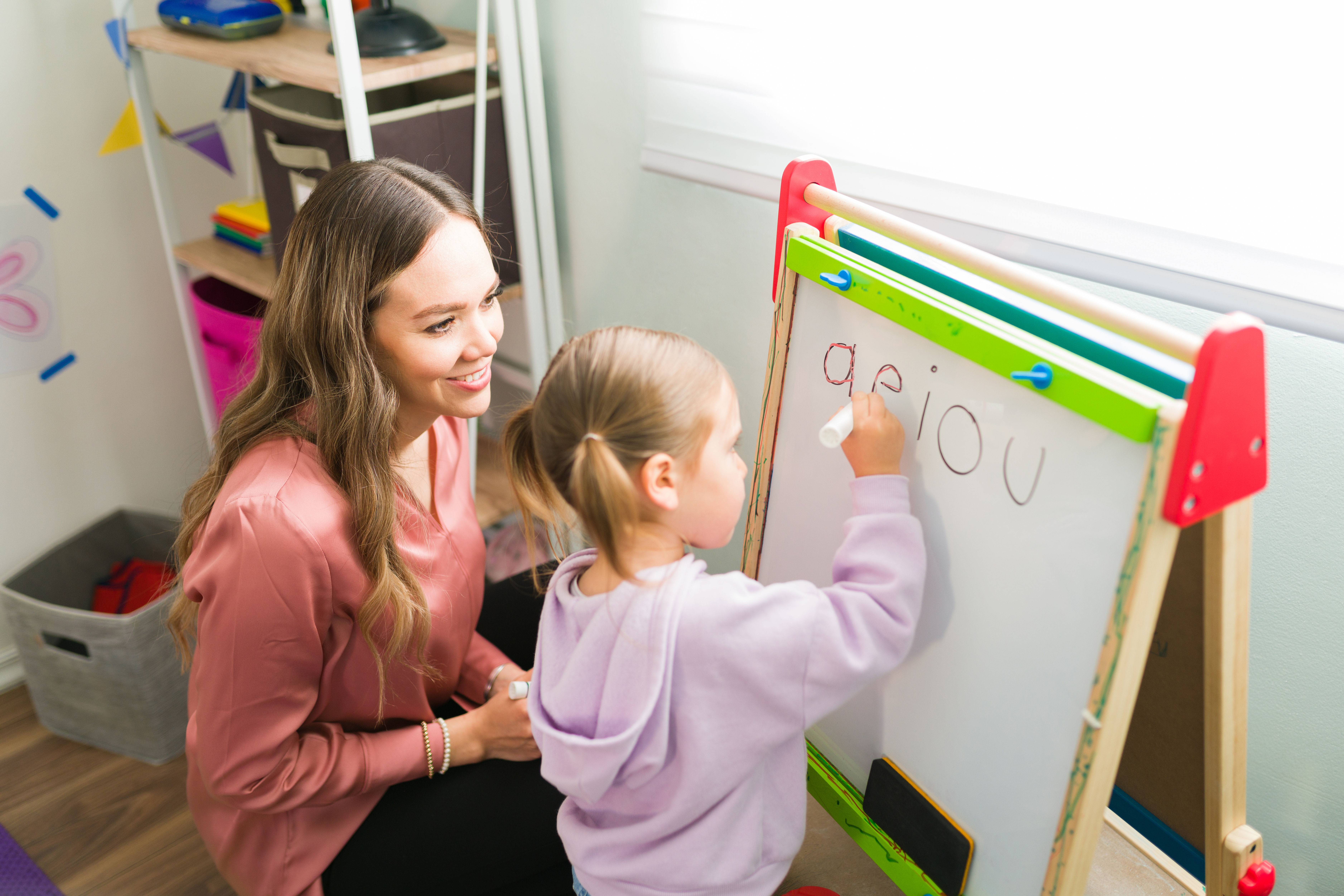 A young child writes letters on a whiteboard easel while an adult kneels beside her and smiles. The classroom setting includes colorful learning materials, storage shelves and children’s supplies, suggesting an early childhood education or teaching activity focused on handwriting and guided learning.