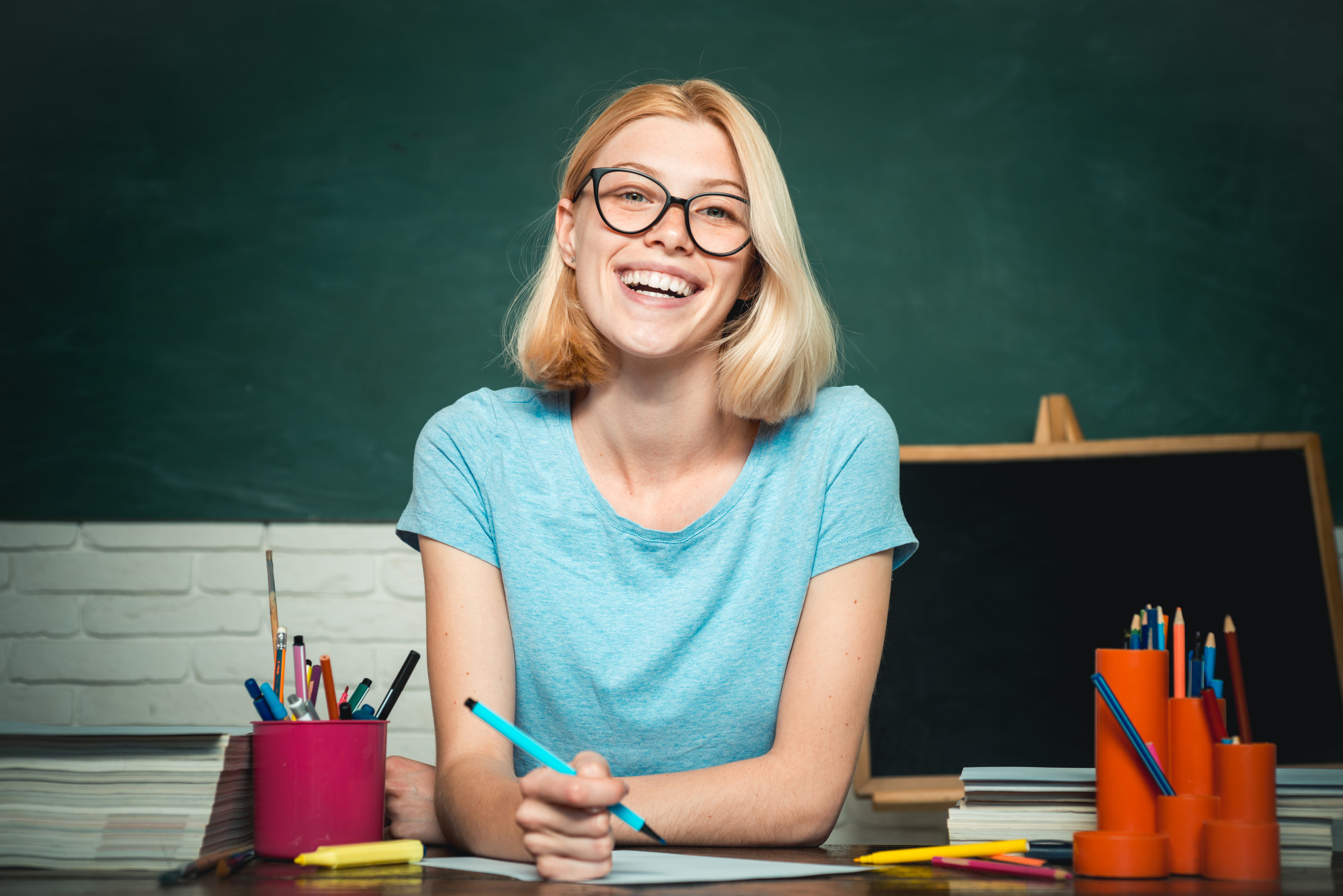 A smiling student sits at a classroom table with a pencil in hand, surrounded by colorful pens, pencils and stacks of paper. A large chalkboard fills the background, and a small blank chalkboard and classroom supplies are visible nearby, suggesting an education or teaching-focused learning environment.