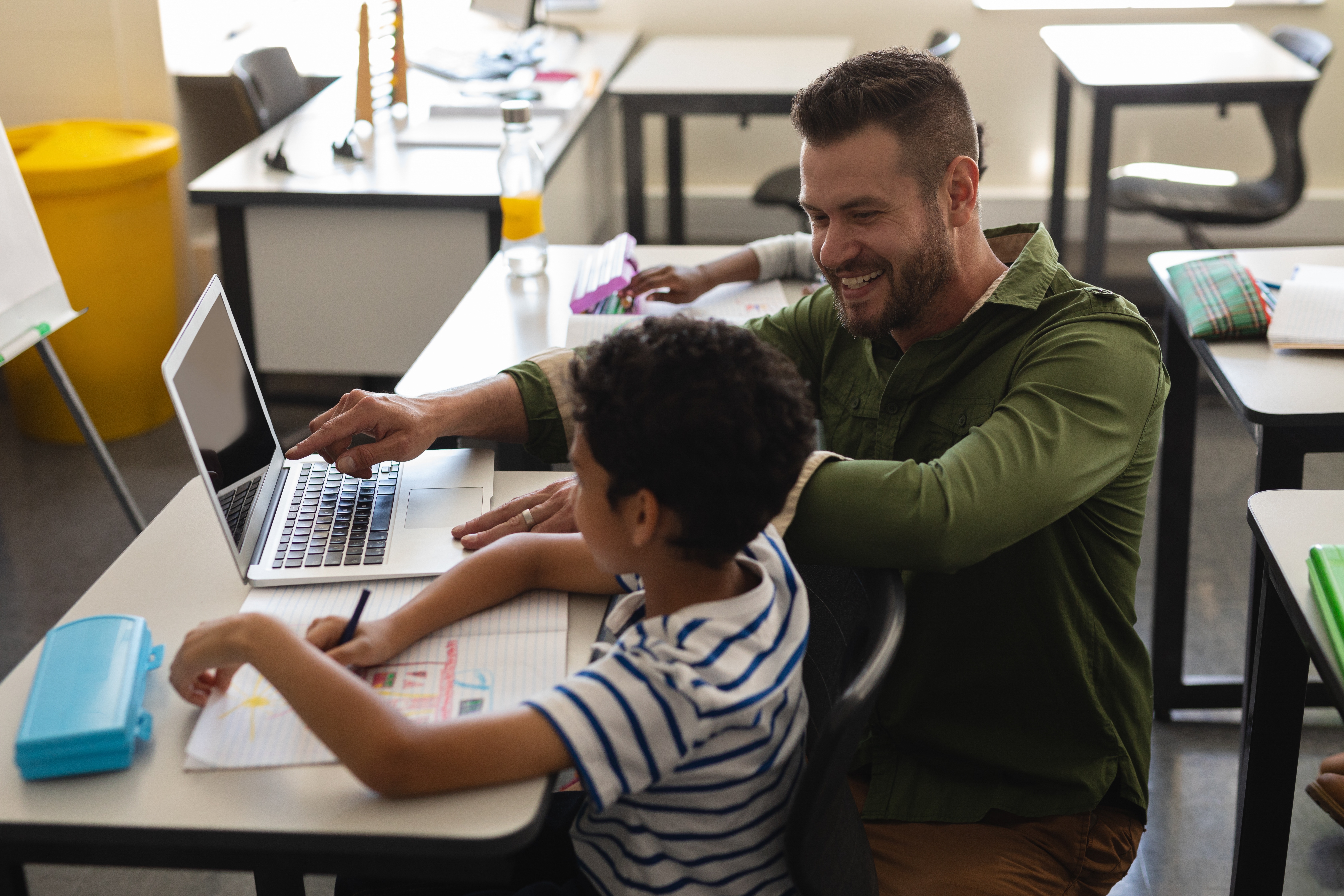 A teacher smiles while helping a young student work on a laptop in a classroom. The student writes in a notebook while the teacher points to the computer screen, suggesting one-on-one instruction or technology-supported learning. Desks, notebooks and classroom supplies are visible in the background.