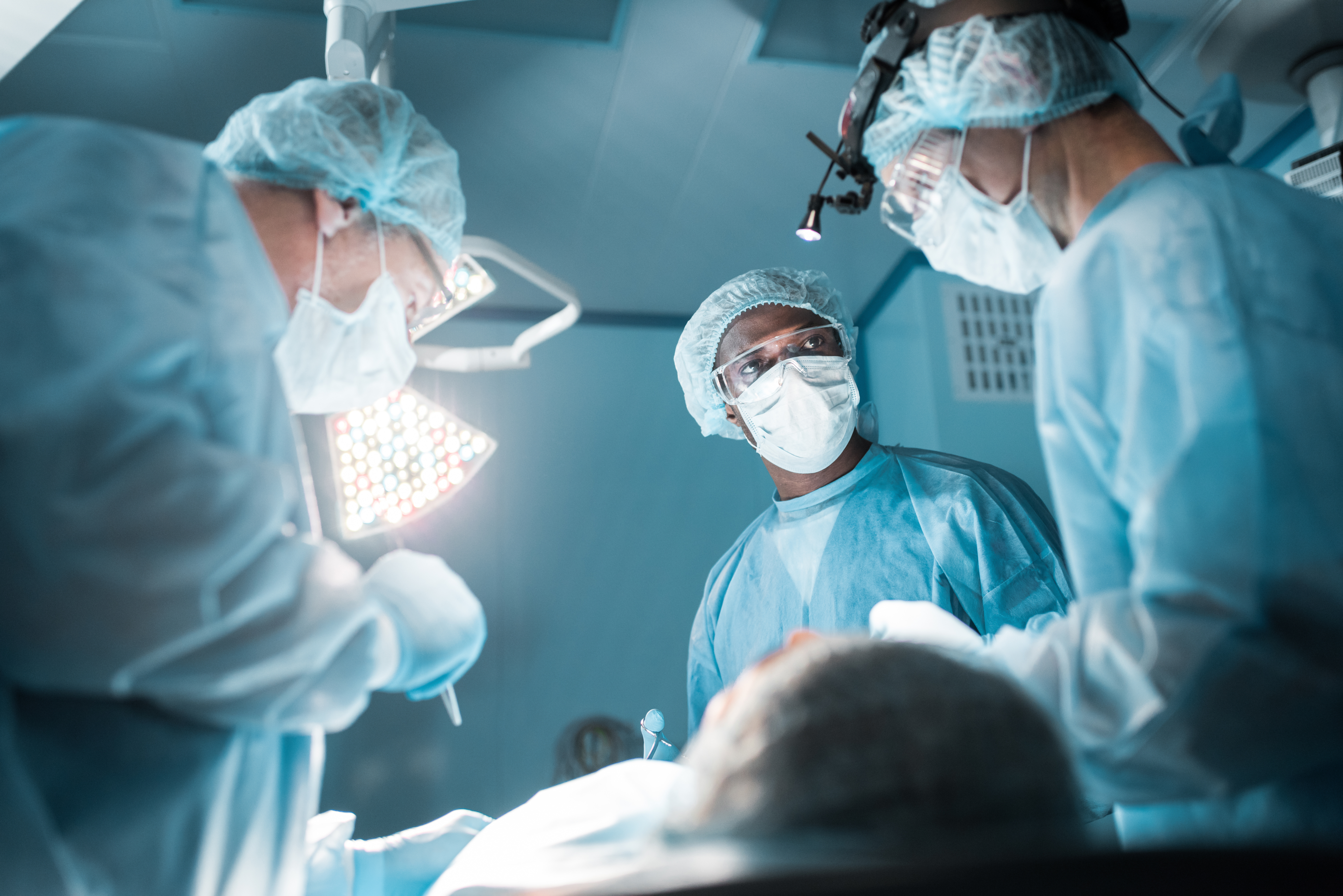 A surgical team wearing masks, hair covers, protective eyewear and sterile gowns works under bright operating room lights. Three medical professionals are gathered around a patient on an operating table, with one team member looking across the surgical field while others focus on the procedure.