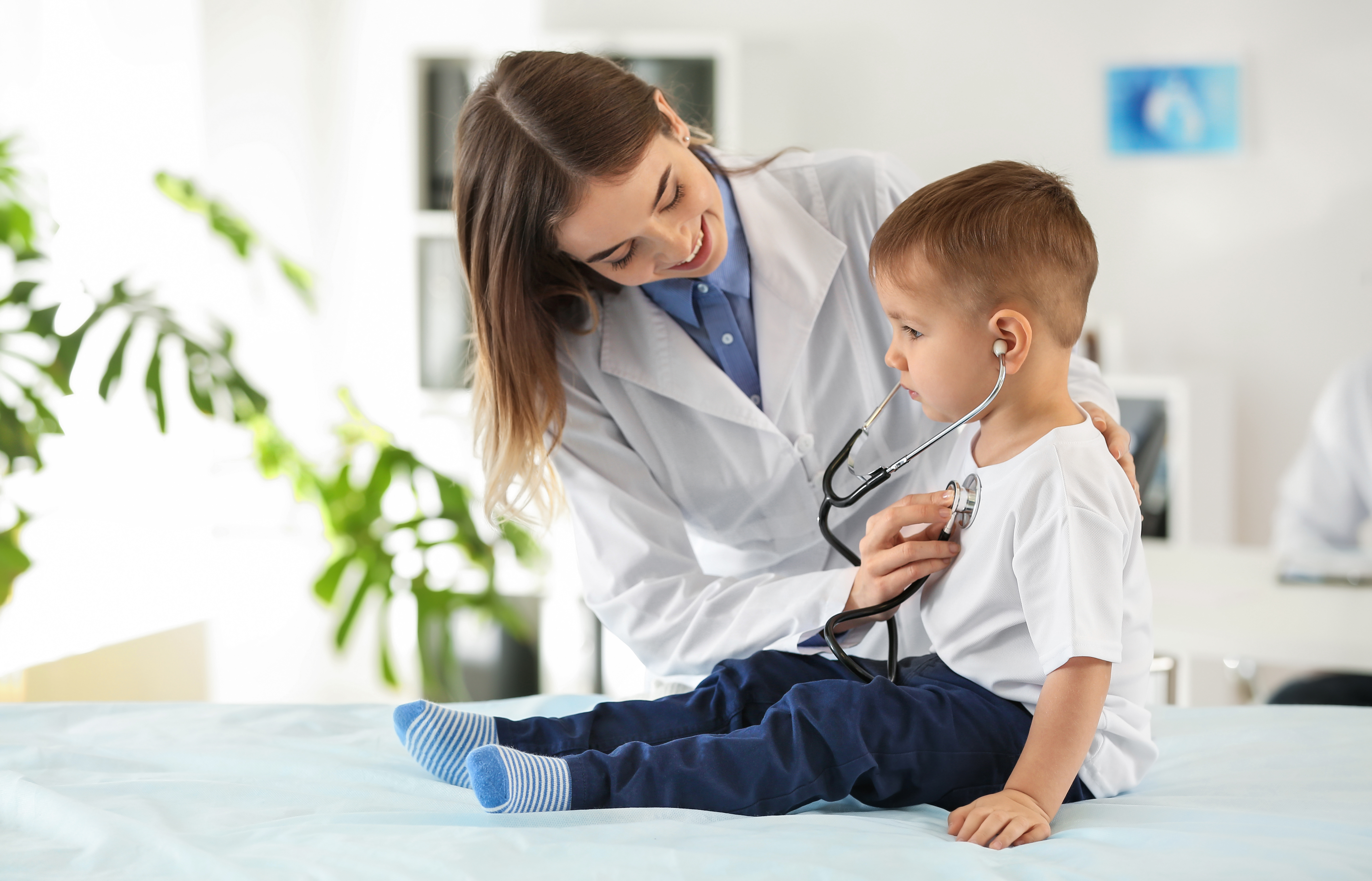 A health care provider in a white coat uses a stethoscope to listen to a young child’s chest during an exam. The child sits on an exam table with the stethoscope earpieces in place, while the provider smiles and guides the checkup in a bright clinical setting.