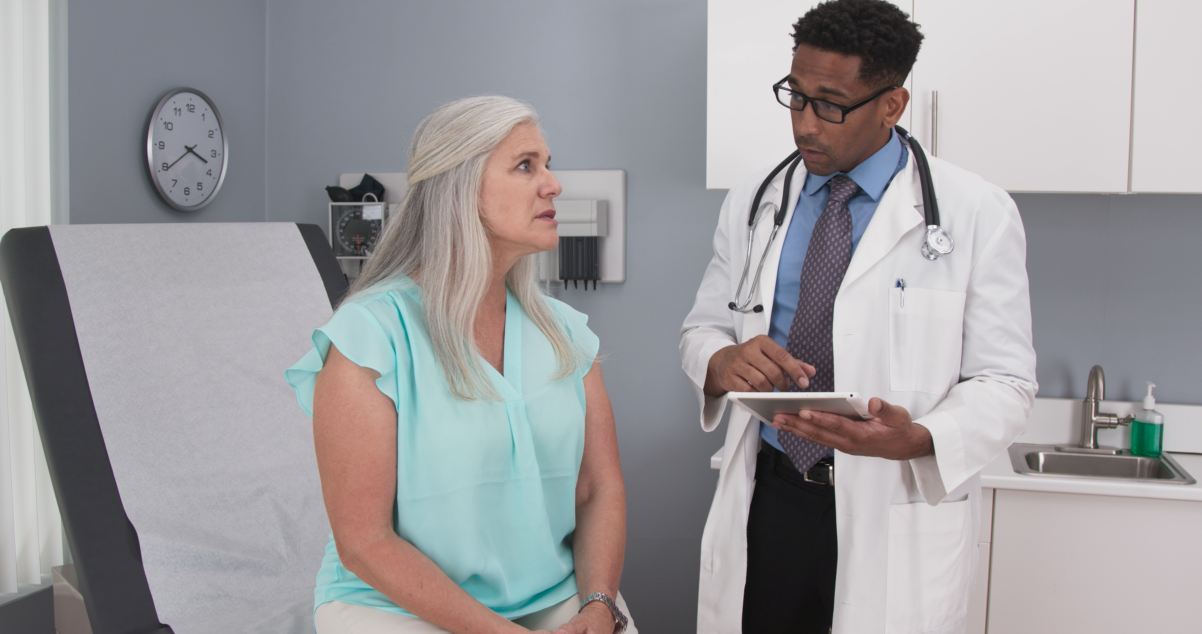 A doctor in a white coat speaks with a patient seated on an exam table in a medical office. The doctor holds a tablet and wears a stethoscope while appearing to discuss care or test results. The patient looks up at the doctor attentively. An exam table, sink, cabinets and medical equipment are visible in the room.