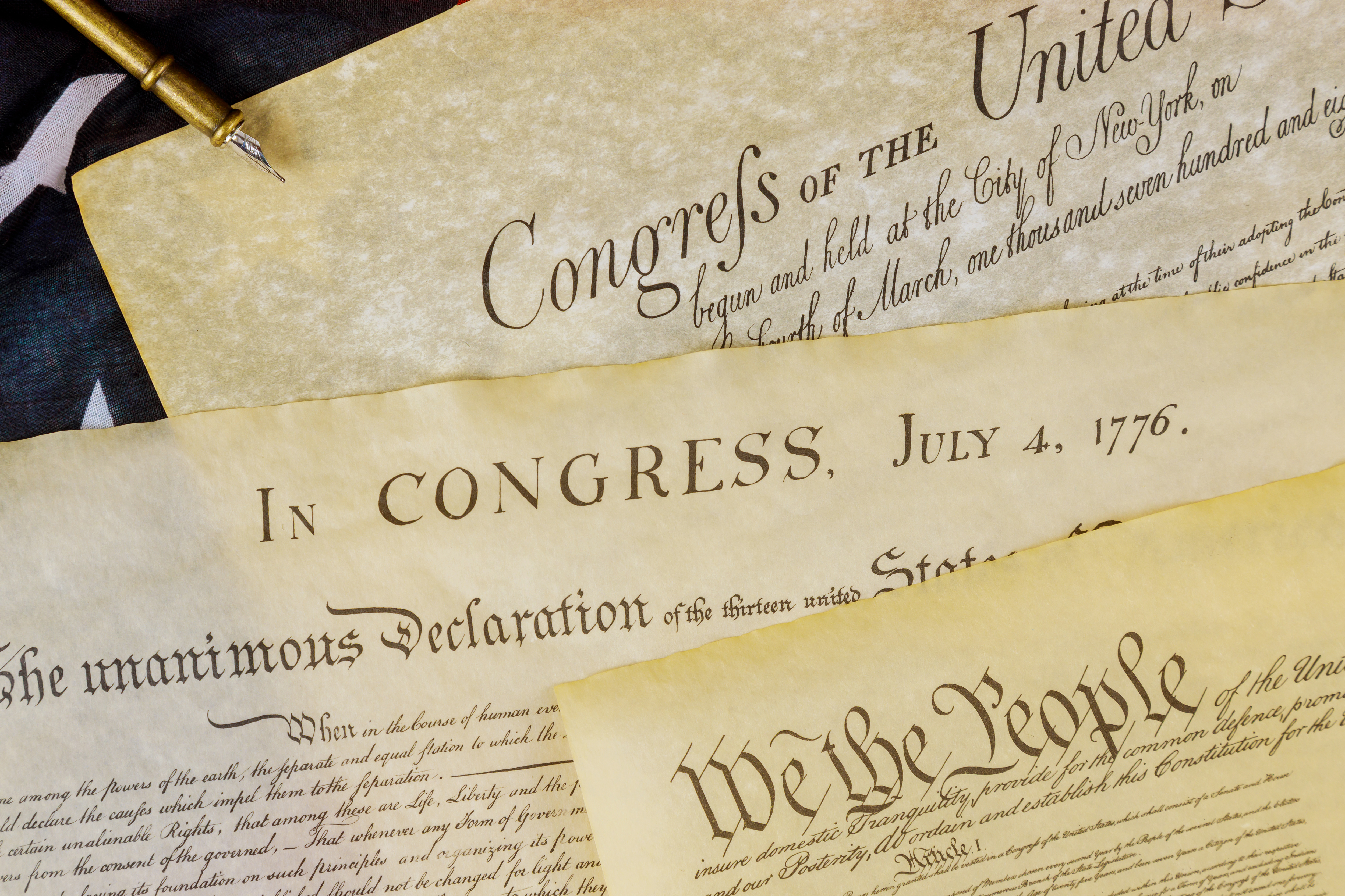 A close-up image shows historical U.S. founding documents layered together, including the Declaration of Independence and U.S. Constitution. Portions of text such as “In Congress, July 4, 1776” and “We the People” are visible, along with part of an American flag and a quill pen, suggesting a humanities, history or government studies theme.