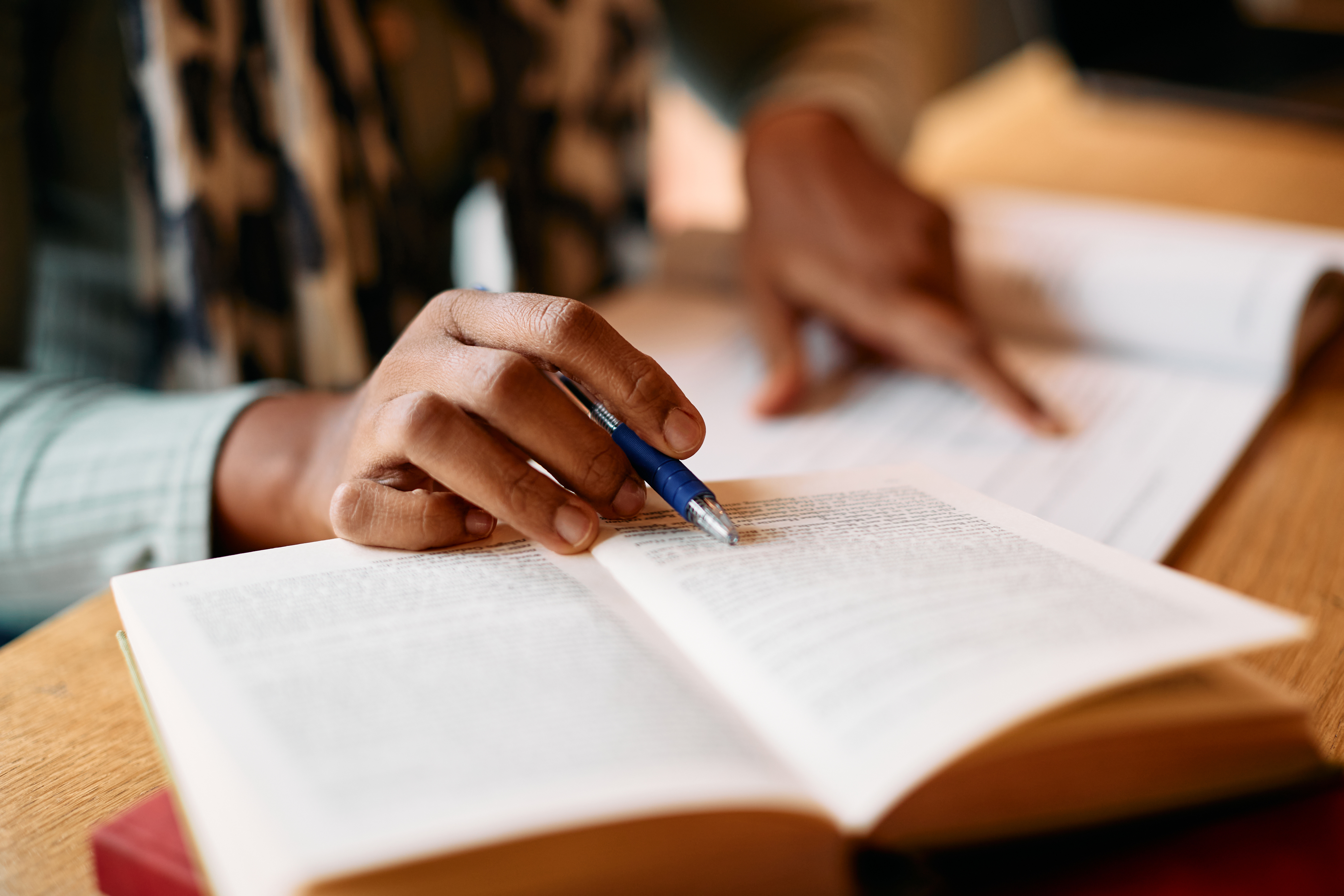 A close-up image shows a person studying at a table, holding a pen over an open book while referencing notes or paperwork nearby. The shallow focus highlights the hand, pen and text on the page, suggesting reading, research, writing or humanities coursework.