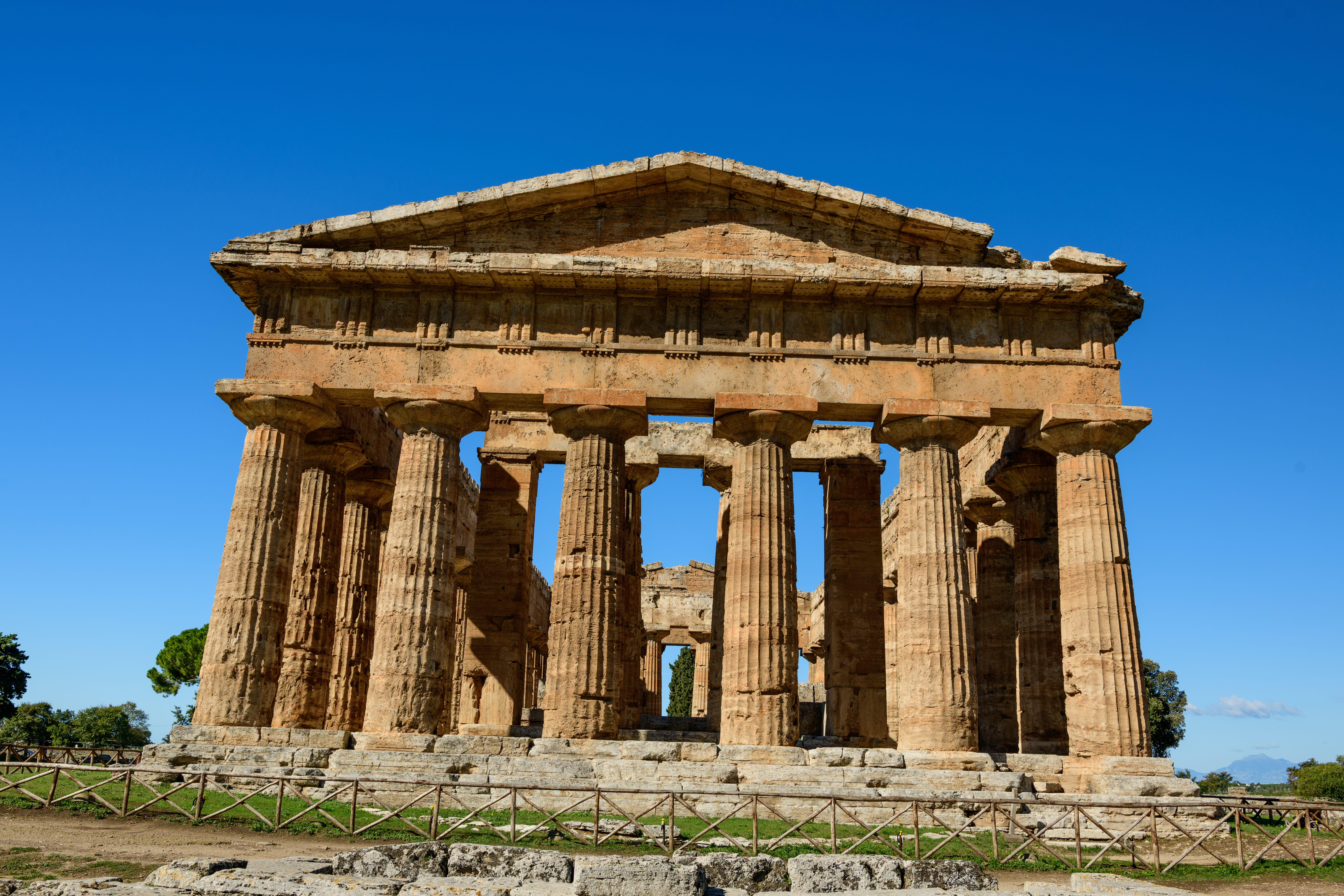 An ancient stone temple with large columns and a triangular roof stands under a bright blue sky. The weathered structure is surrounded by low fencing, with stone steps and ruins visible in front. The image suggests a humanities, history, archaeology or world civilizations theme.