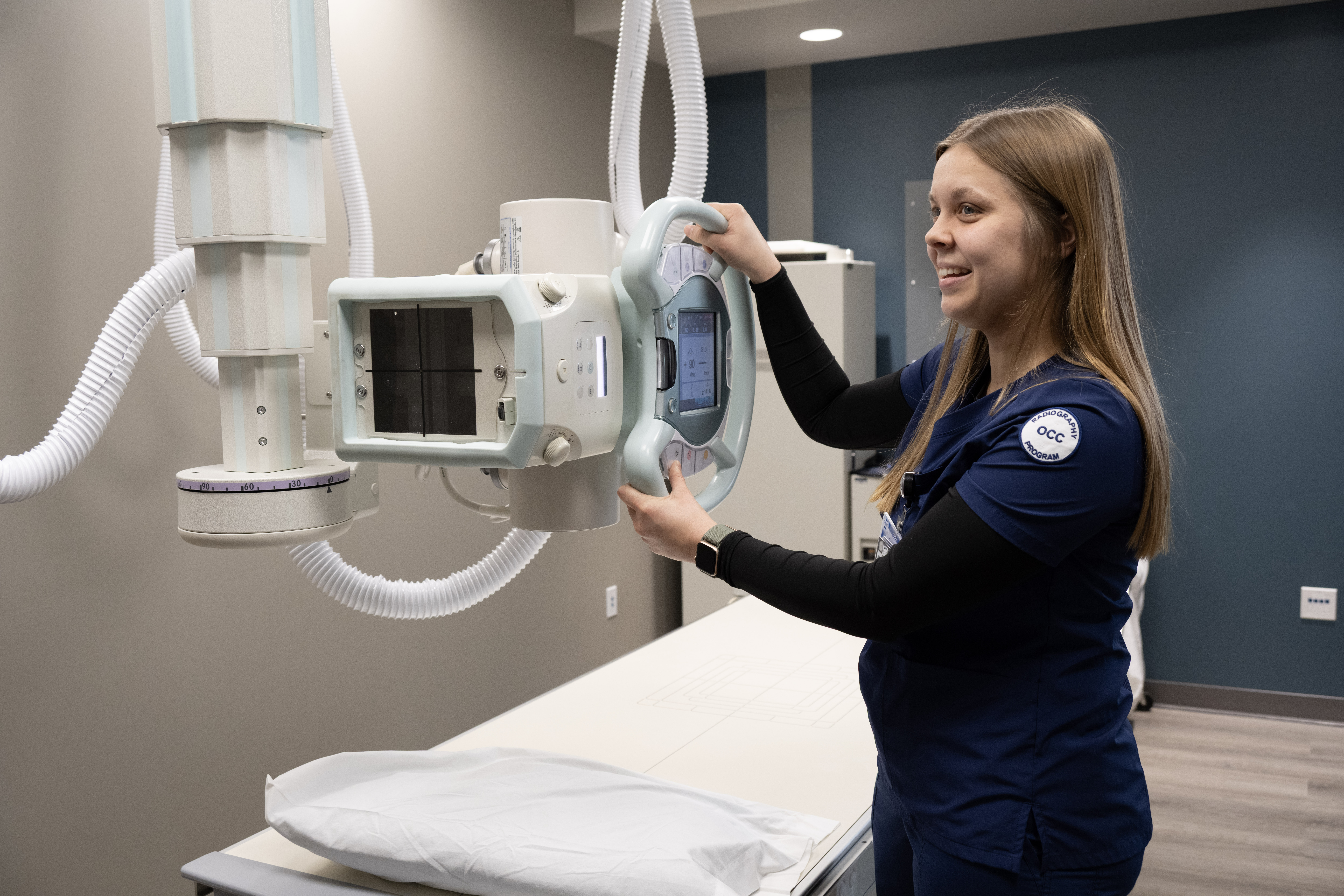 Radiography student positions and adjusts an X-ray imaging unit above an examination table in the Health Professions Center lab.