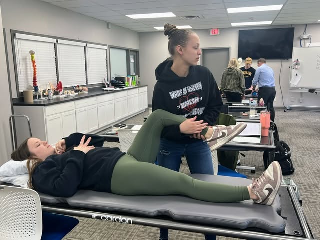 Two students practice a rehabilitative therapy technique in a classroom lab. One student lies on a treatment table while another stands beside her and supports her raised leg, appearing to guide a stretch or range-of-motion exercise. Other students, treatment tables and lab equipment are visible in the background.