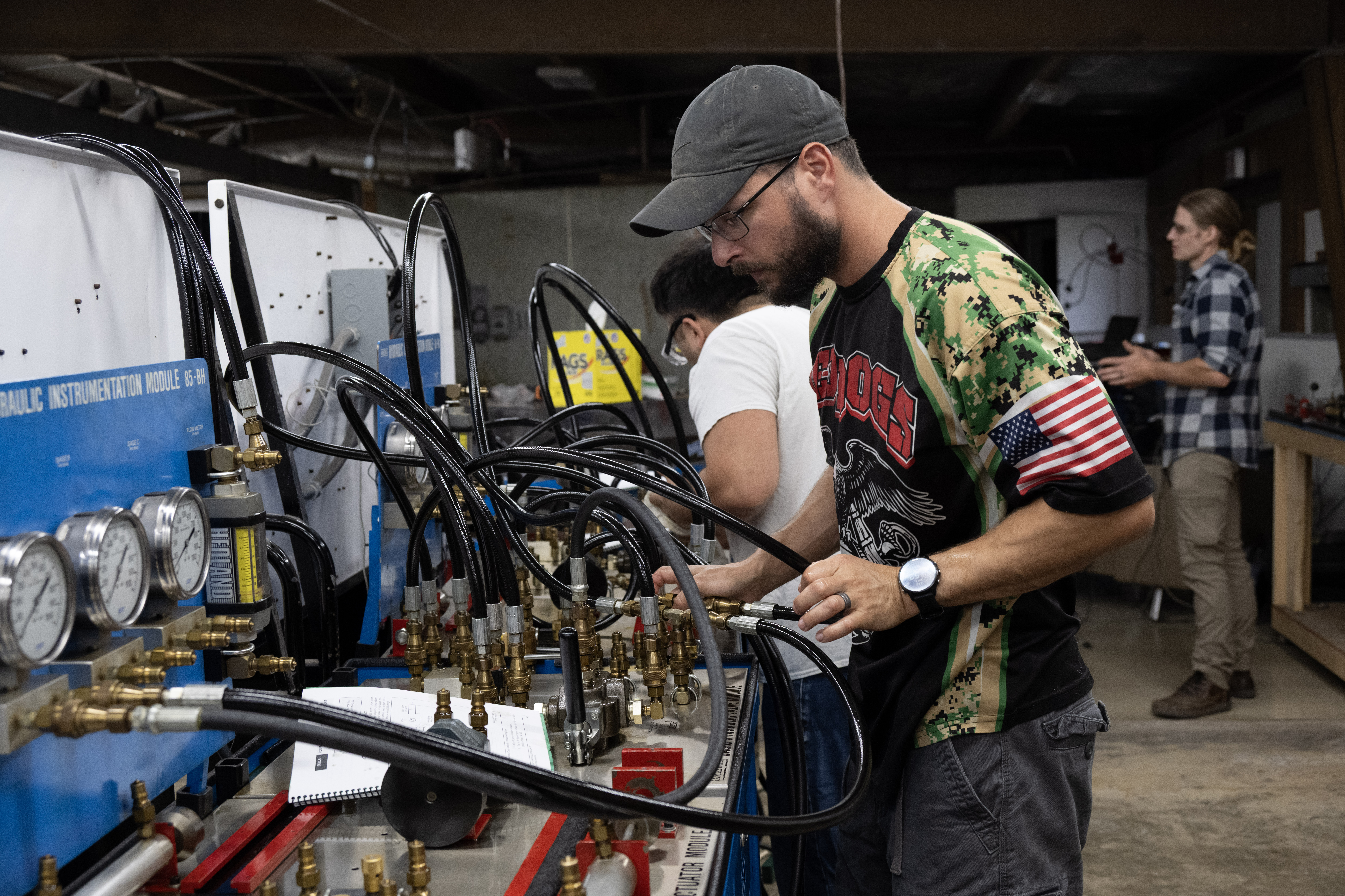 Students work with hydraulic training equipment in an industrial maintenance lab, connecting hoses and monitoring guages.