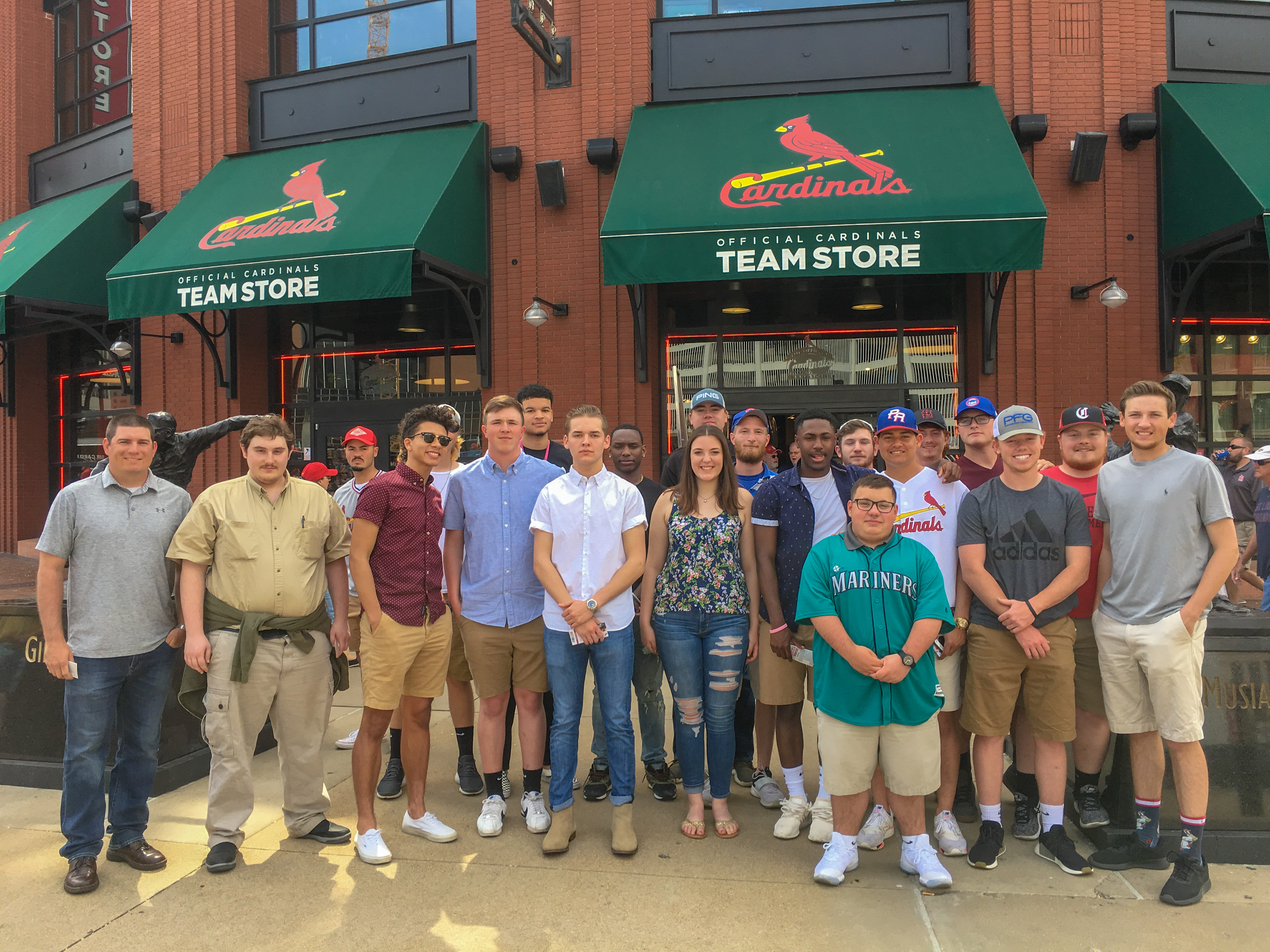 A group of sport management students poses outside the Official Cardinals Team Store during a field trip to Busch Stadium. The students stand together on the sidewalk in front of the brick stadium building and green awnings with the Cardinals logo. Some wear Cardinals or other baseball-themed shirts and caps, while others wear casual clothes. The group photo captures the students gathered closely together during the visit.