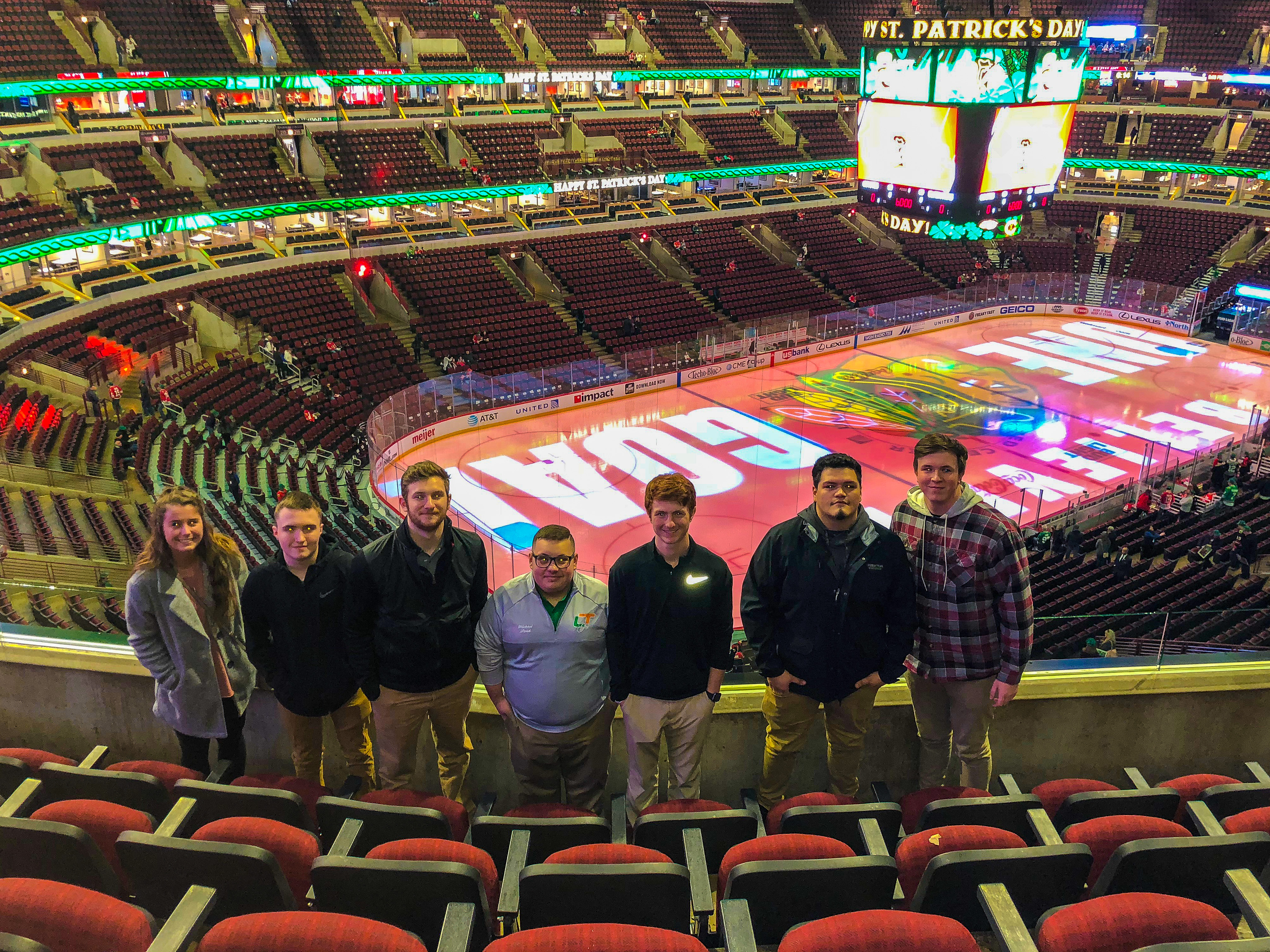 A group of sport management students stands in the seating area at the United Center during a field trip. The arena’s ice surface is lit with colorful graphics, and the scoreboard and ribbon boards display St. Patrick’s Day-themed messages. Rows of red seats, the rink, arena signage and scattered spectators are visible behind the students.