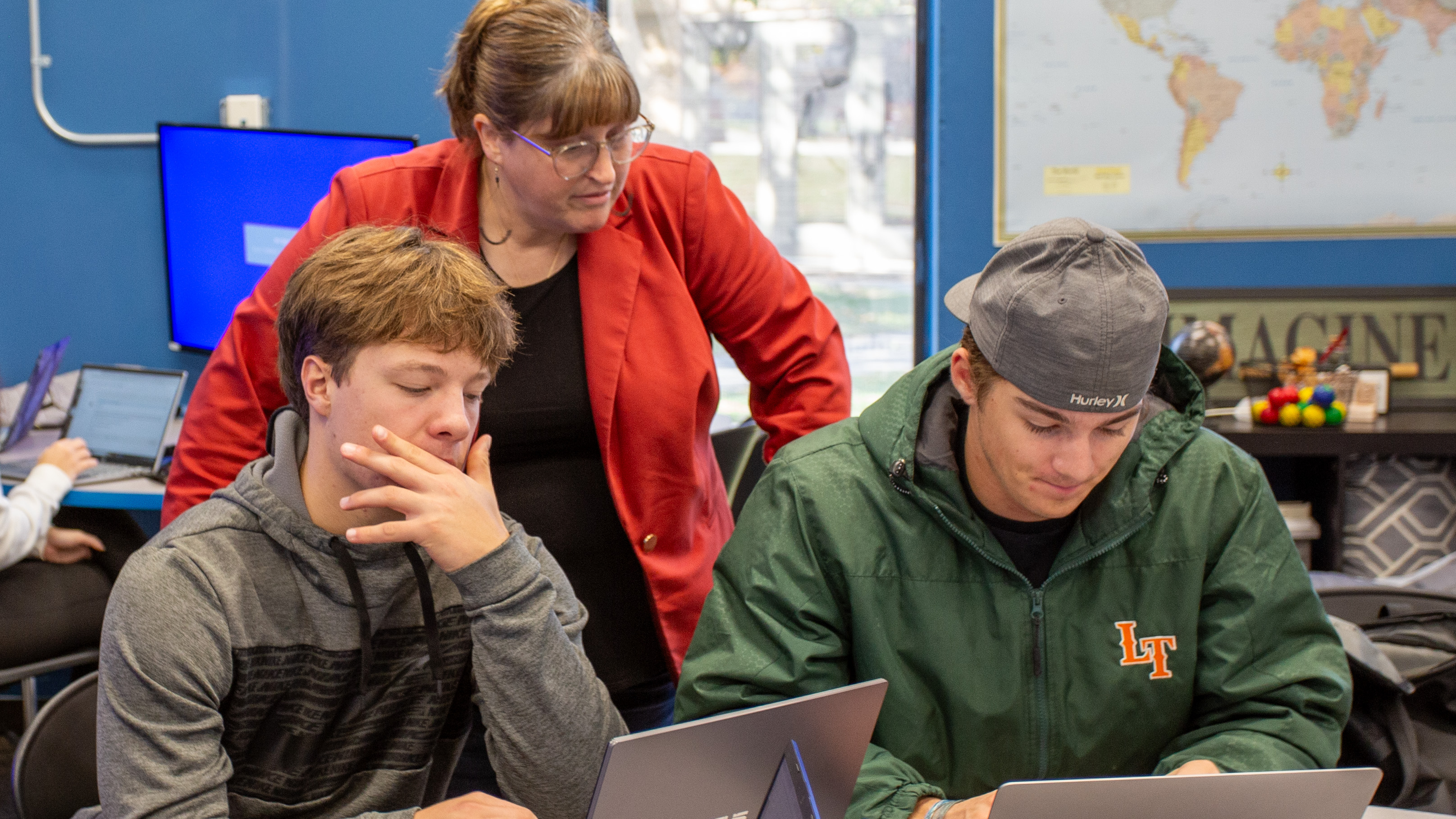 A marketing instructor stands behind two students as they work on laptops in a classroom. One student looks at his screen thoughtfully while another types beside him. A world map, classroom decorations and another student working at a computer are visible in the background, suggesting a hands-on marketing or business class activity.