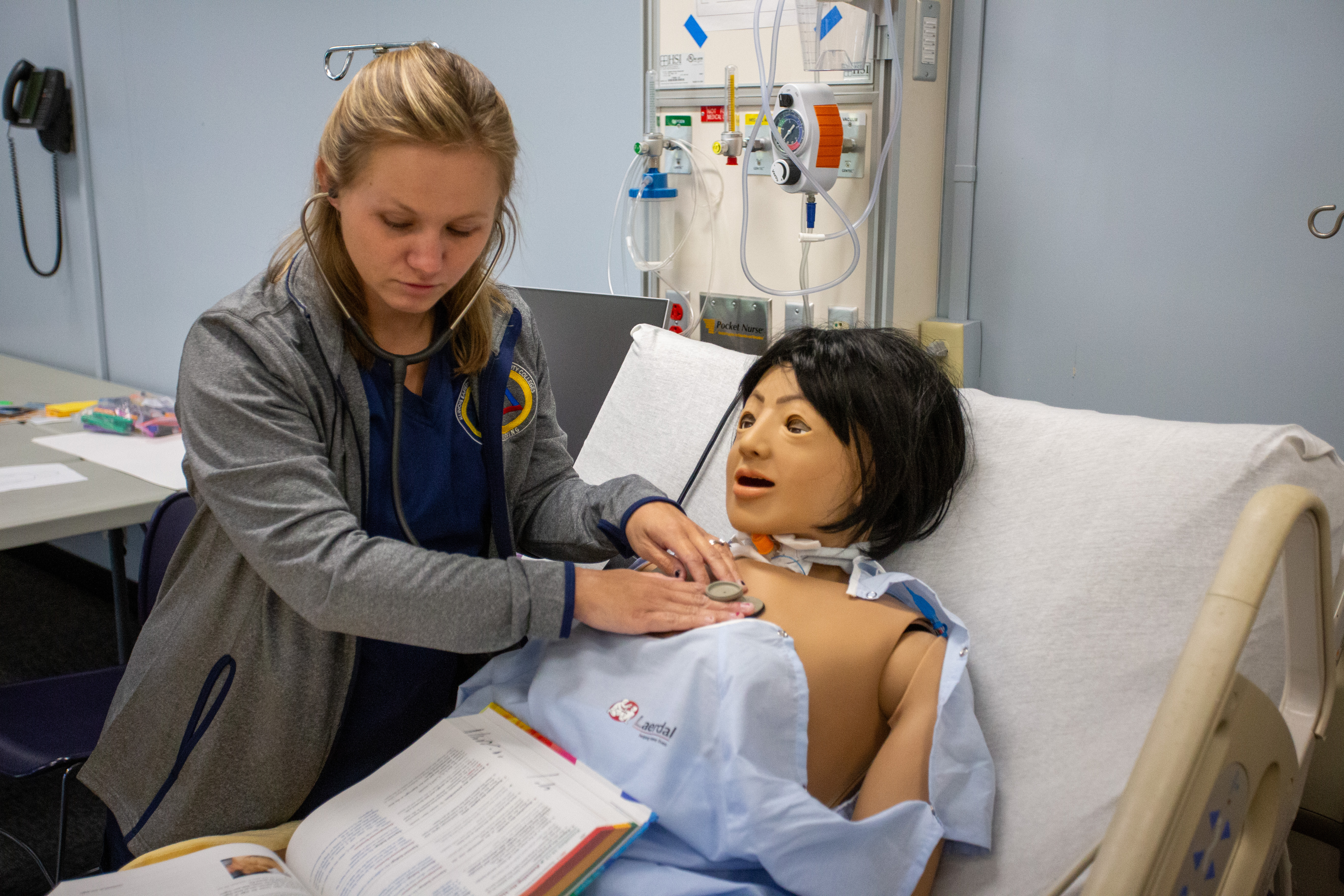 A nursing student uses a stethoscope to assess a medical training mannequin lying in a hospital bed during a lab exercise. The student places the stethoscope on the mannequin’s chest while reviewing an open textbook or workbook nearby. Medical gas outlets, tubing and classroom lab equipment are visible behind the bed, suggesting a hands-on nursing skills practice setting.