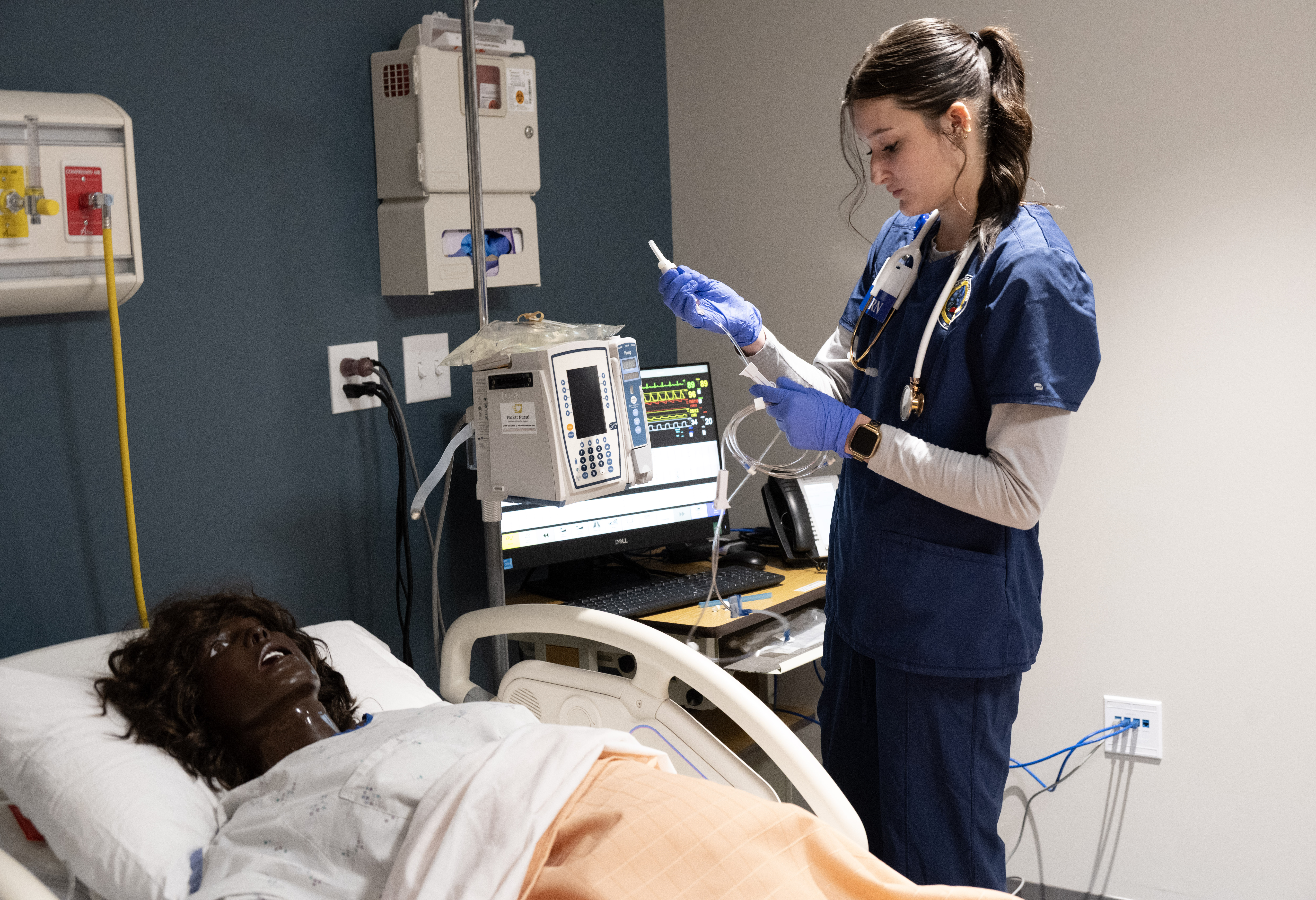 Nursing student in a clinical simulation lab prepares IV tubing beside a patient-care manikin in a hospital bed, with monitoring equipment nearby.