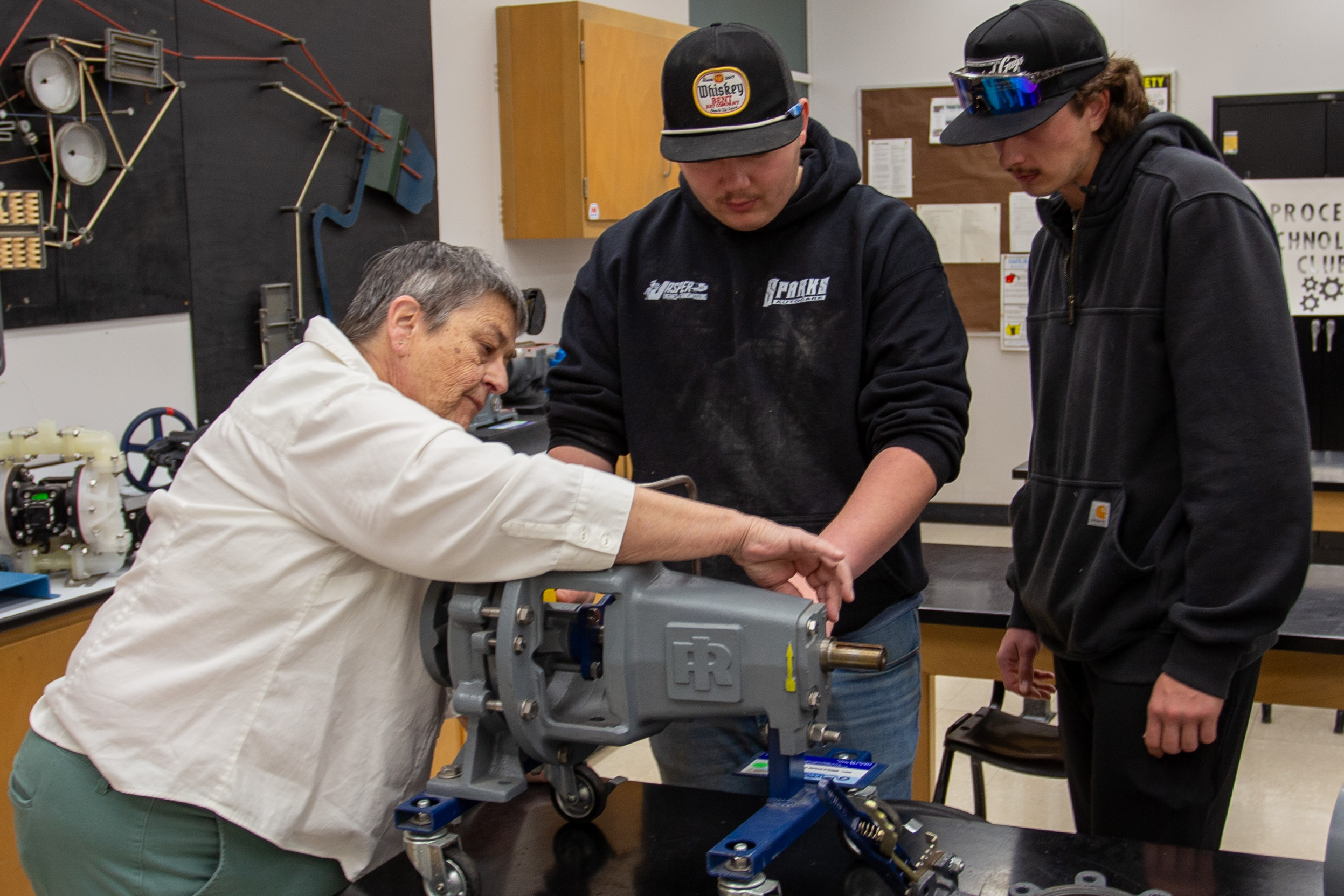 Instructor Tina Lindley demonstrates a process technology training pump or mechanical component to two students in a lab. Lindley points to the equipment while the students watch closely. Worktables, classroom equipment, diagrams and process technology materials are visible in the background, suggesting hands-on instruction with industrial systems and mechanical troubleshooting.