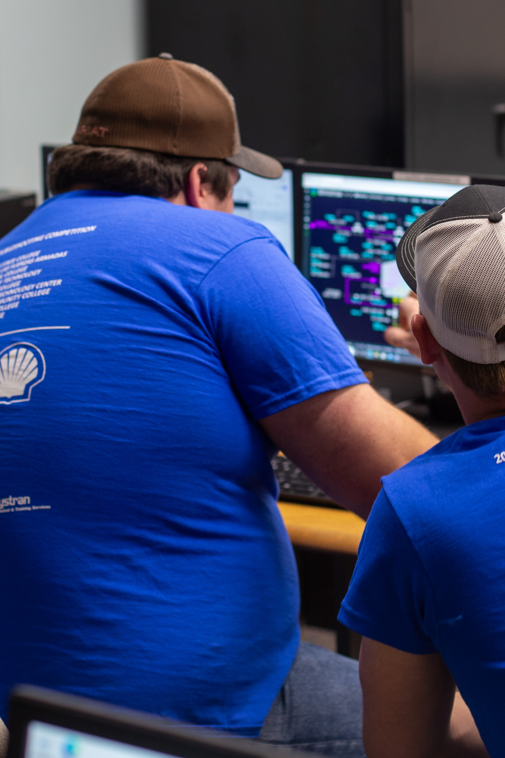 Two process technology students sit together at a computer workstation while reviewing a colorful technical diagram or process control display on a monitor. One student points toward the screen as they discuss the information. Both students wear blue industry training shirts, and additional monitors and classroom technology are visible around them.