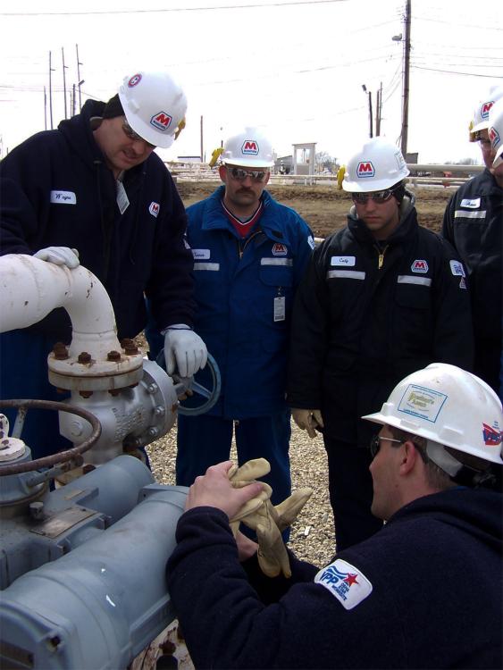 A group of process technology students and instructors wearing hard hats and safety glasses gather around industrial piping and valve equipment at an outdoor training site. One person operates or points to a valve while others watch closely. The setting includes large pipes, utility equipment and worksite infrastructure, suggesting hands-on training with industrial systems and field safety procedures.