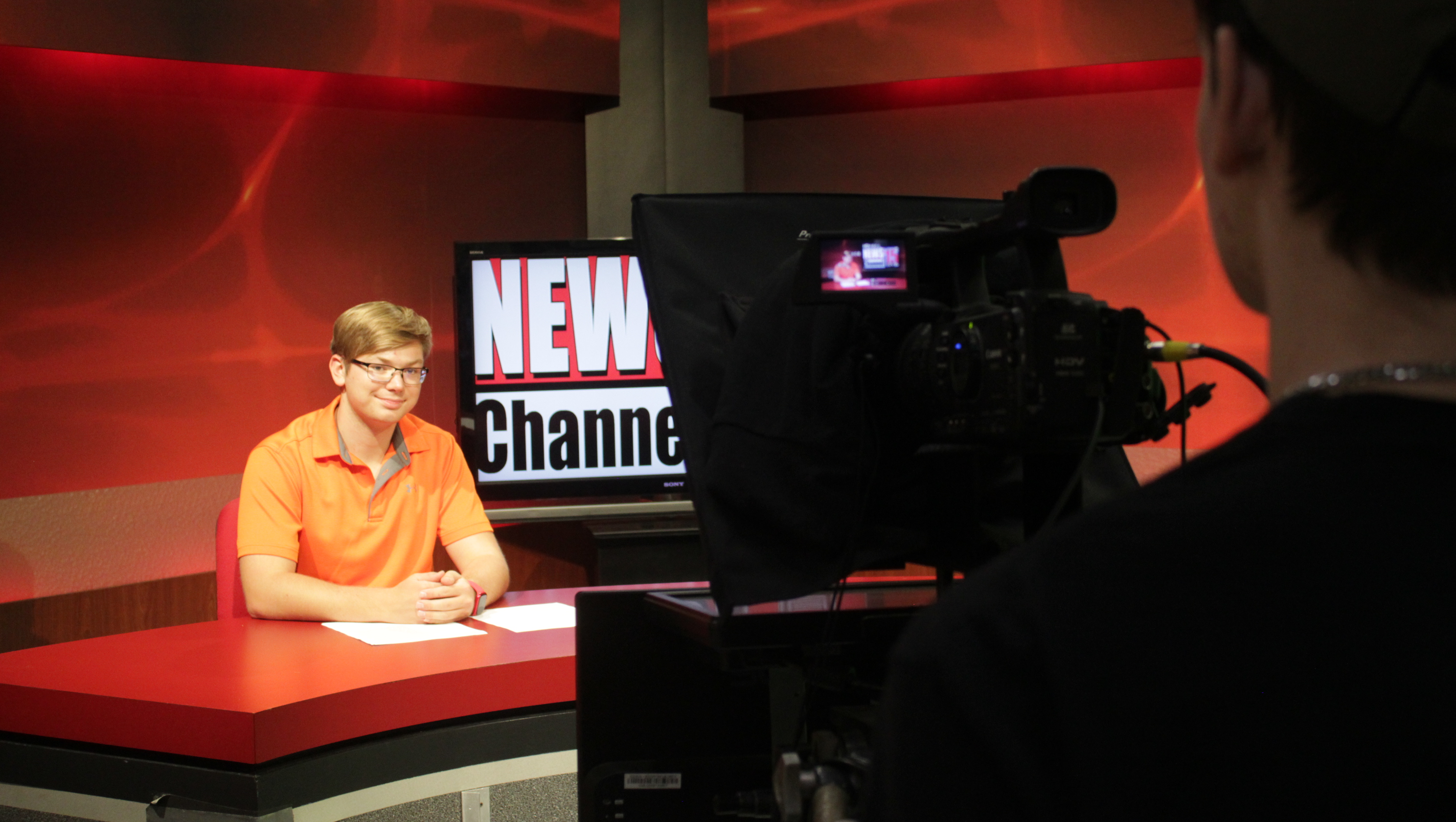 A student sits behind a news desk in a television studio while another student operates a professional video camera in the foreground. A “News Channel 15” display and red broadcast set pieces are visible behind the anchor desk, suggesting hands-on training in radio, television and digital media production.