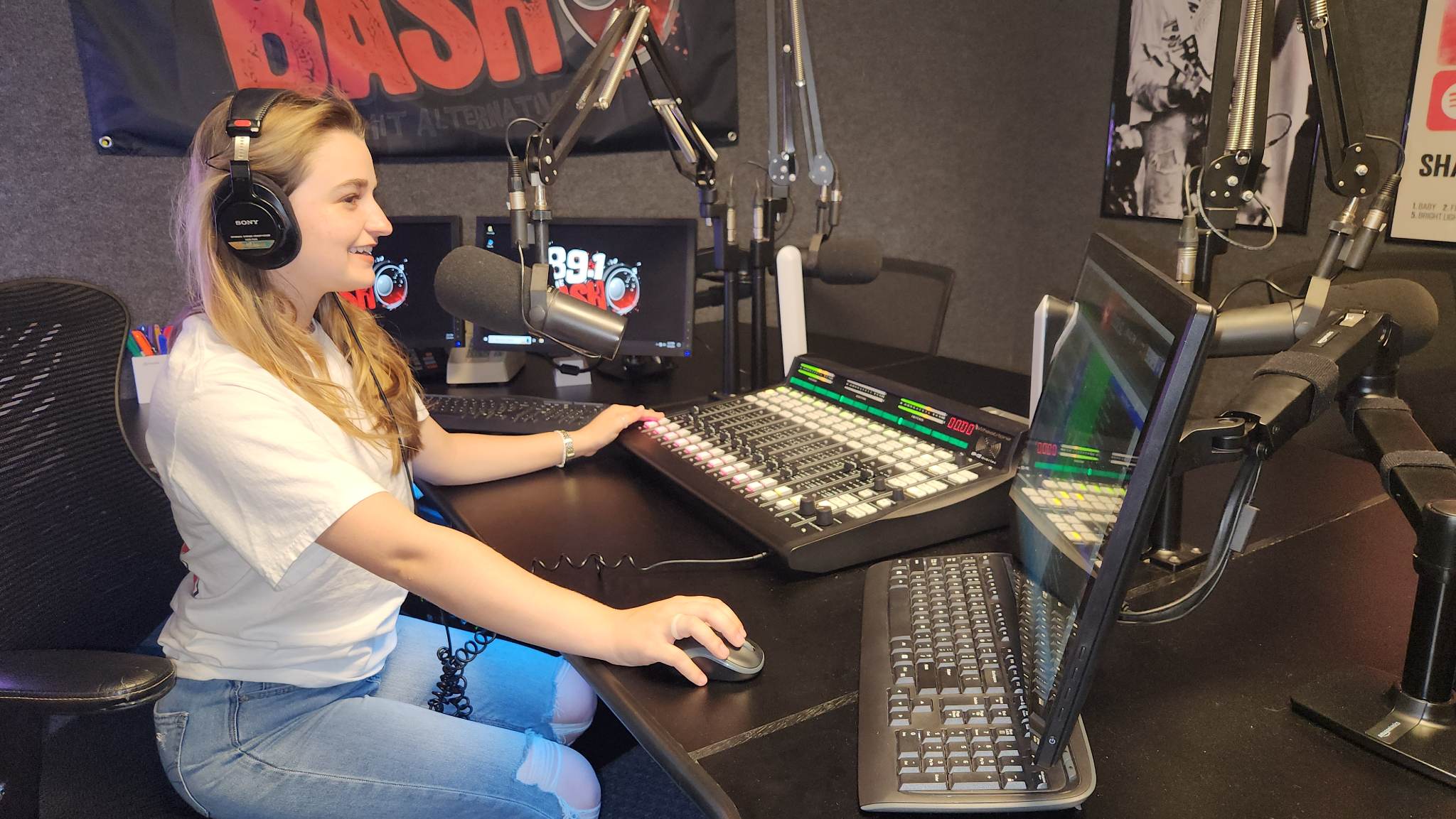 A student wearing headphones works at a radio station control board, using a computer mouse and audio mixer while seated near multiple microphones. Computer screens in the studio display station graphics, suggesting hands-on training in radio, television and digital media production, including audio operation, broadcasting and on-air media work.