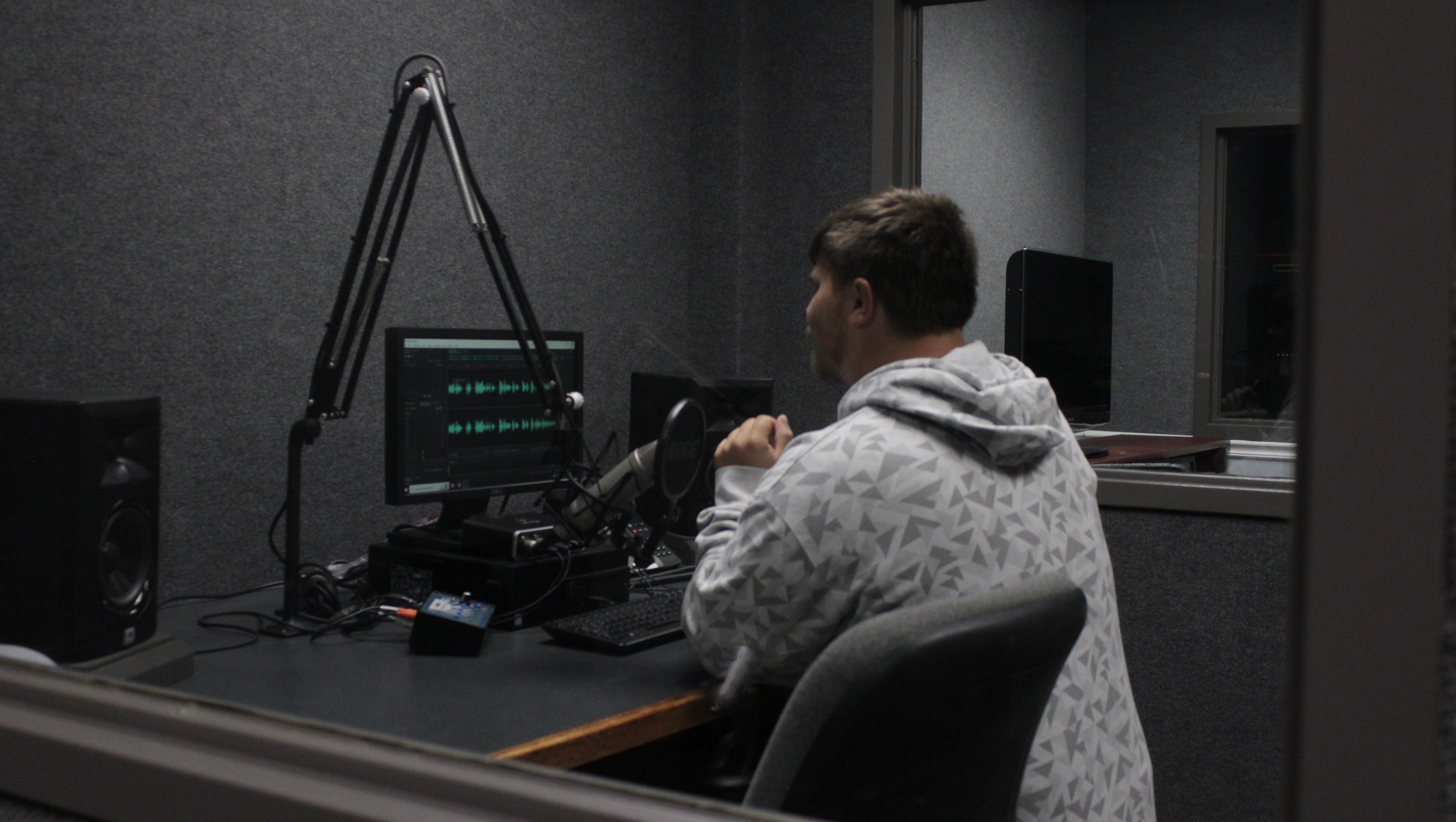 A student sits in a recording booth at a microphone, speaking toward a computer screen displaying audio waveforms. Surrounded by studio equipment, speakers and editing software, the scene highlights hands-on learning in digital media through audio production, recording and content creation.
