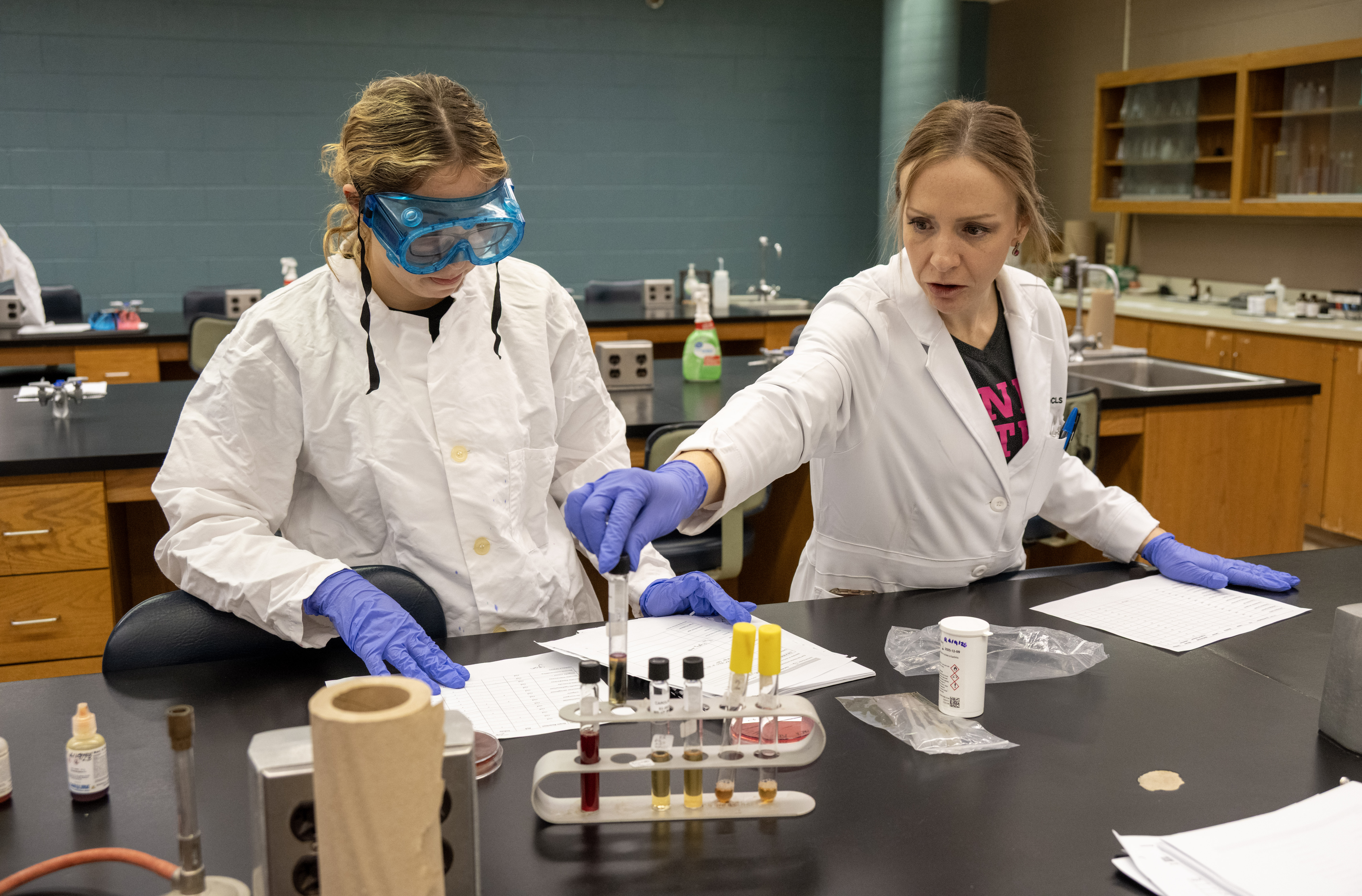Instructor guides a student wearing protective goggles, gloves, and a lab coat during a science experiment. Test tubes, samples, and laboratory supplies are arranged on the lab bench.