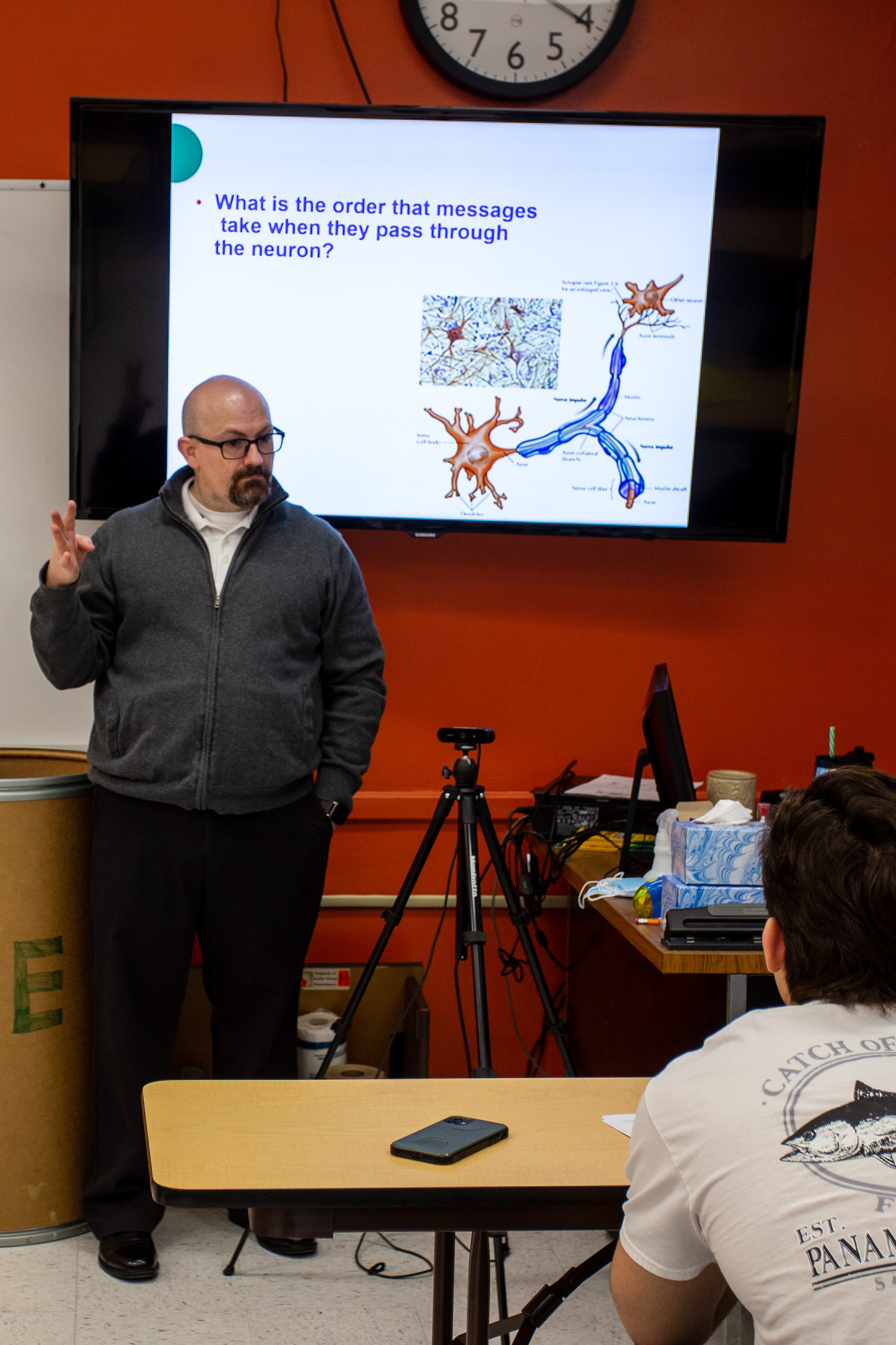 Dr. Phil Thorsen leads a psychology lecture in a classroom while standing beside a screen displaying diagrams of neurons. The slide asks, “What is the order that messages take when they pass through the neuron?” A student watches from a desk in the foreground, and classroom equipment, a tripod and a whiteboard are visible around the room.