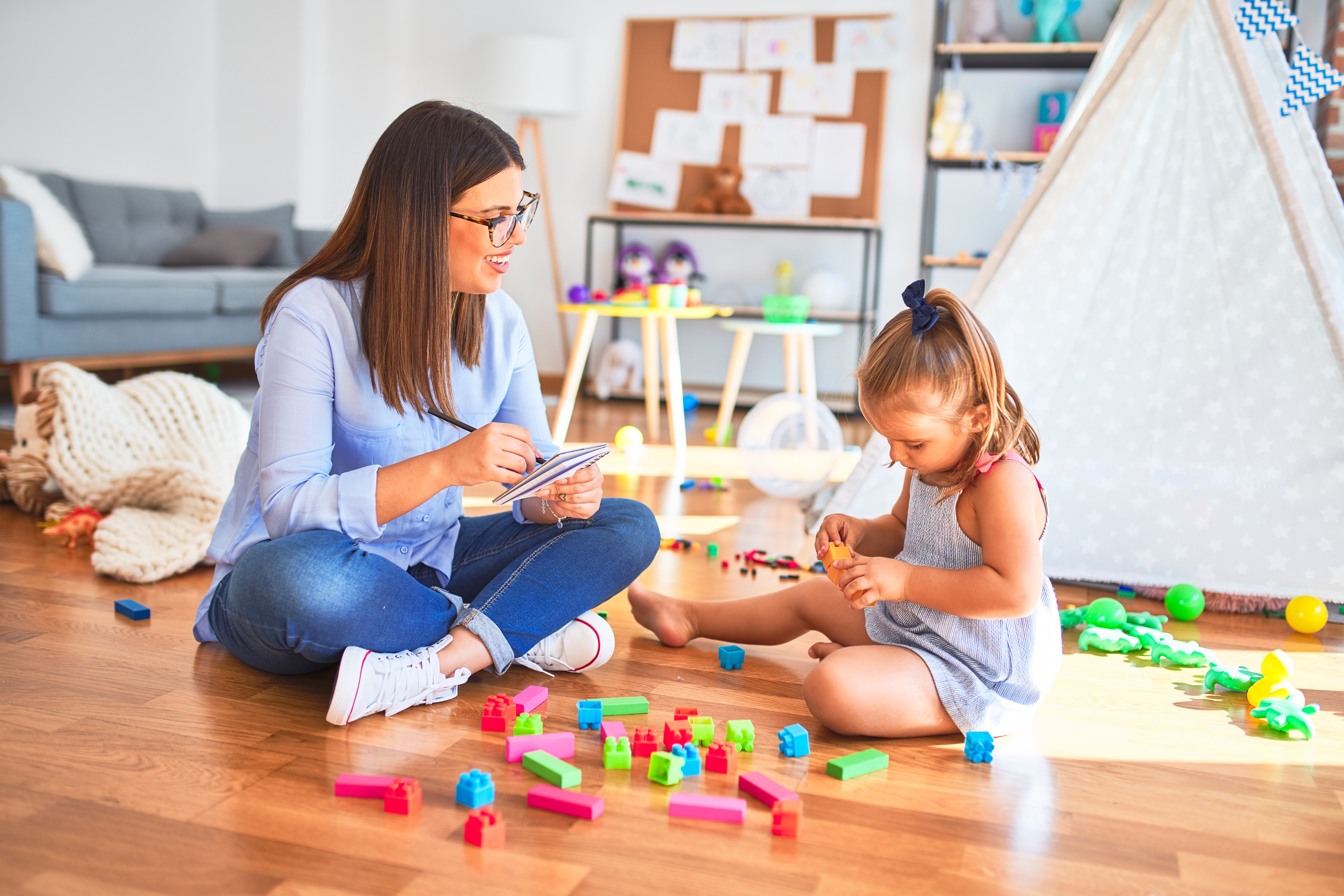 An adult sits with a young child during a play-based activity, taking notes while the child plays with colorful building blocks on the floor. The setting includes toys, a play tent and child-friendly materials, suggesting a social sciences, counseling or child development activity focused on observation, interaction and supportive guidance.