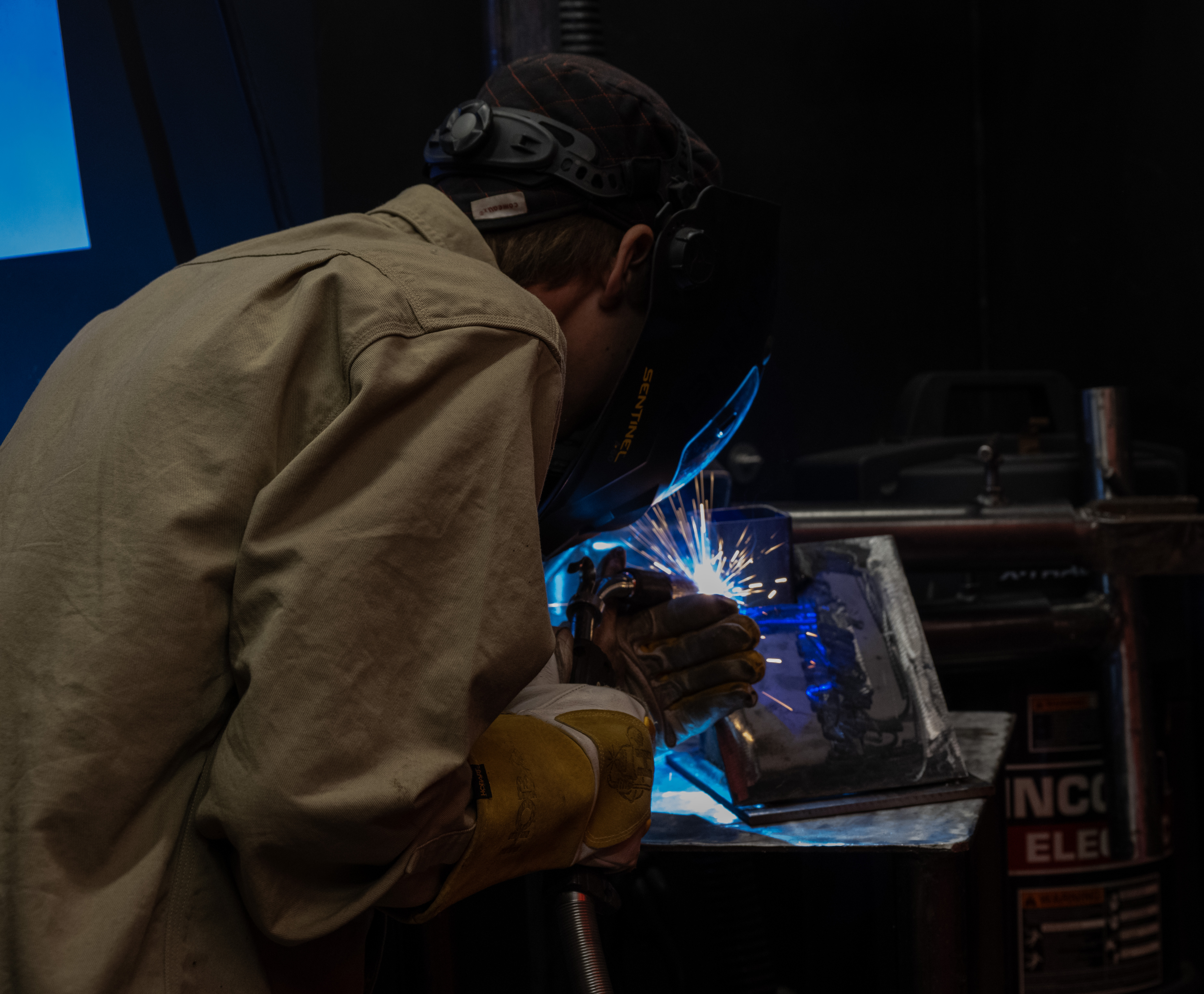 Student performs a welding exercise inside a welding booth, wearing a welding helmet, gloves, and protective jacket as sparks illuminate the metal workpiece.