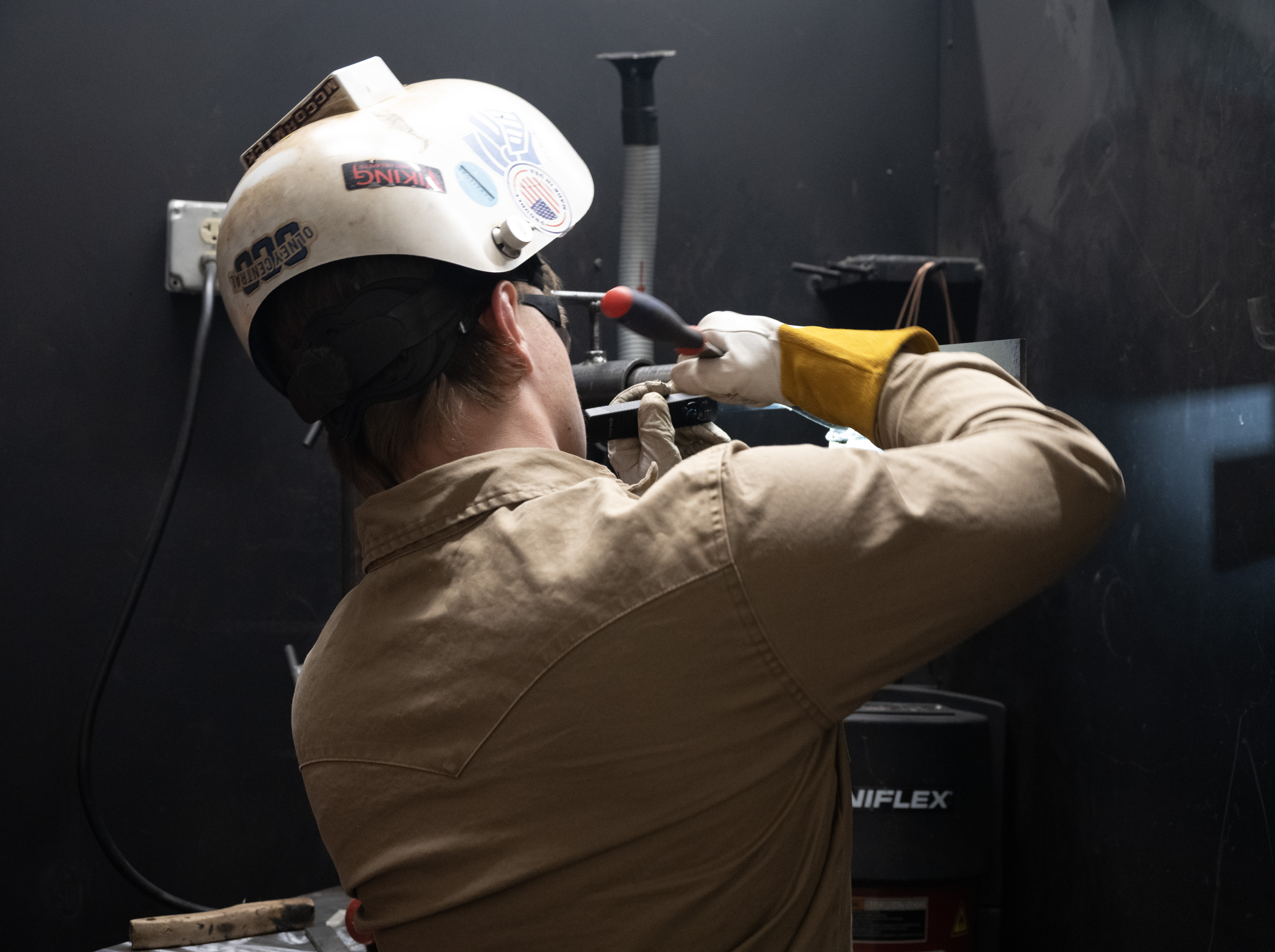 Student practices welding in a training booth while wearing protective safety equipment, during a hands-on fabrication exercise.