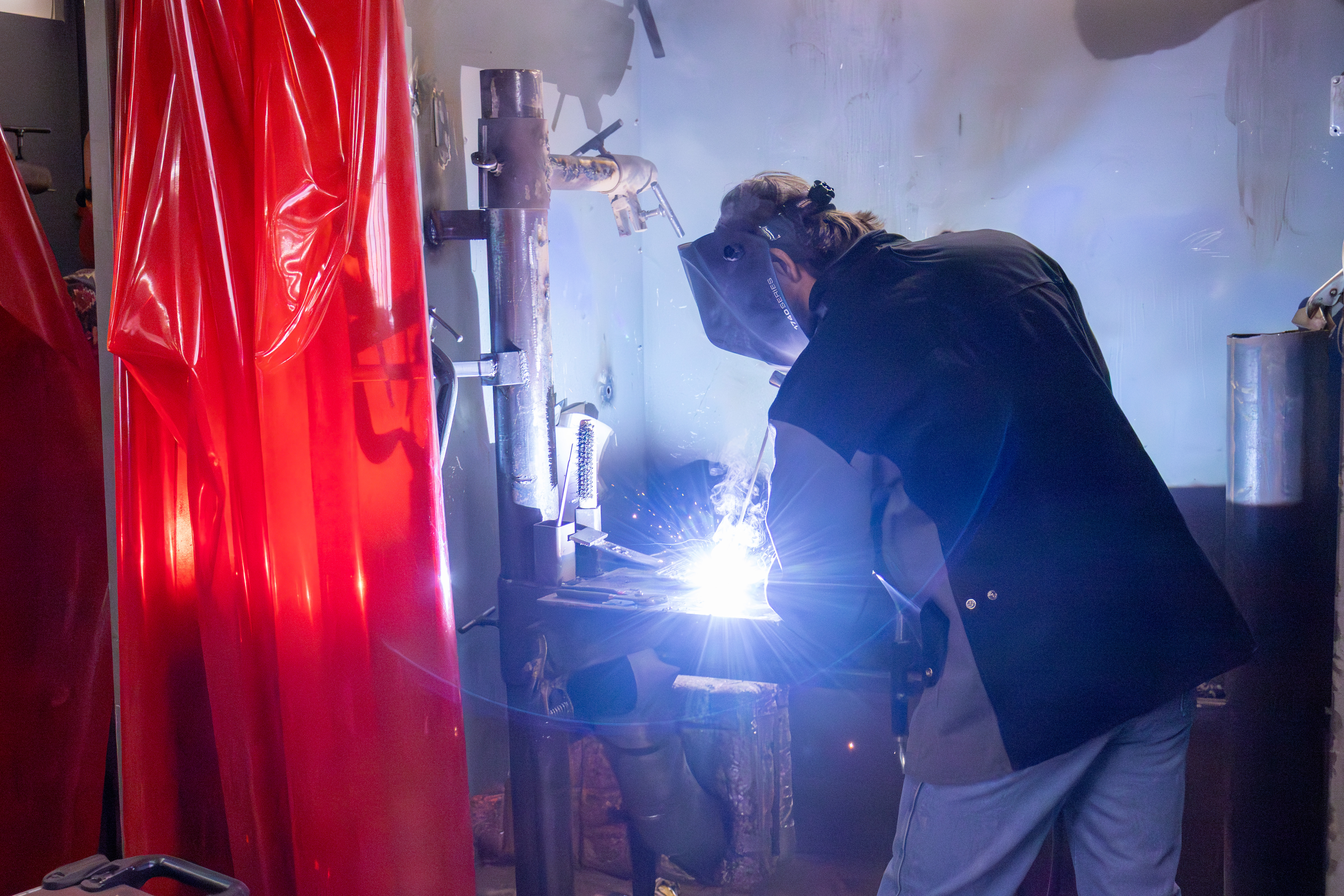 A welding student wearing a protective helmet works inside a welding booth, creating a bright arc and sparks while joining metal components. Red welding curtains hang nearby, and smoke and blue-white light from the weld fill the workspace, suggesting hands-on training in welding and fabrication.
